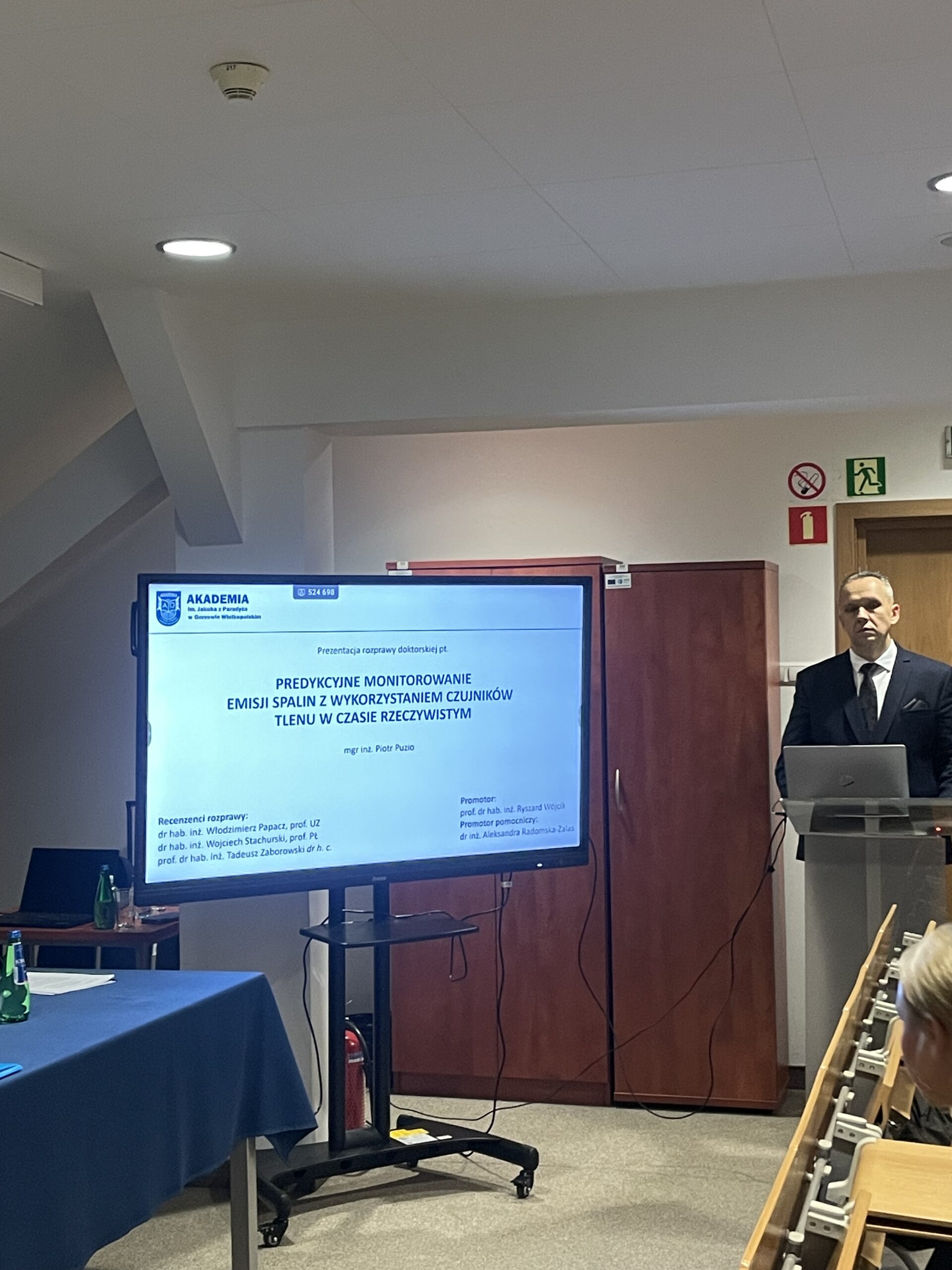 Na pierwszym planie monitor z wyświetlanym tytułem obrony. Obok przy mównicy stoi doktorant.