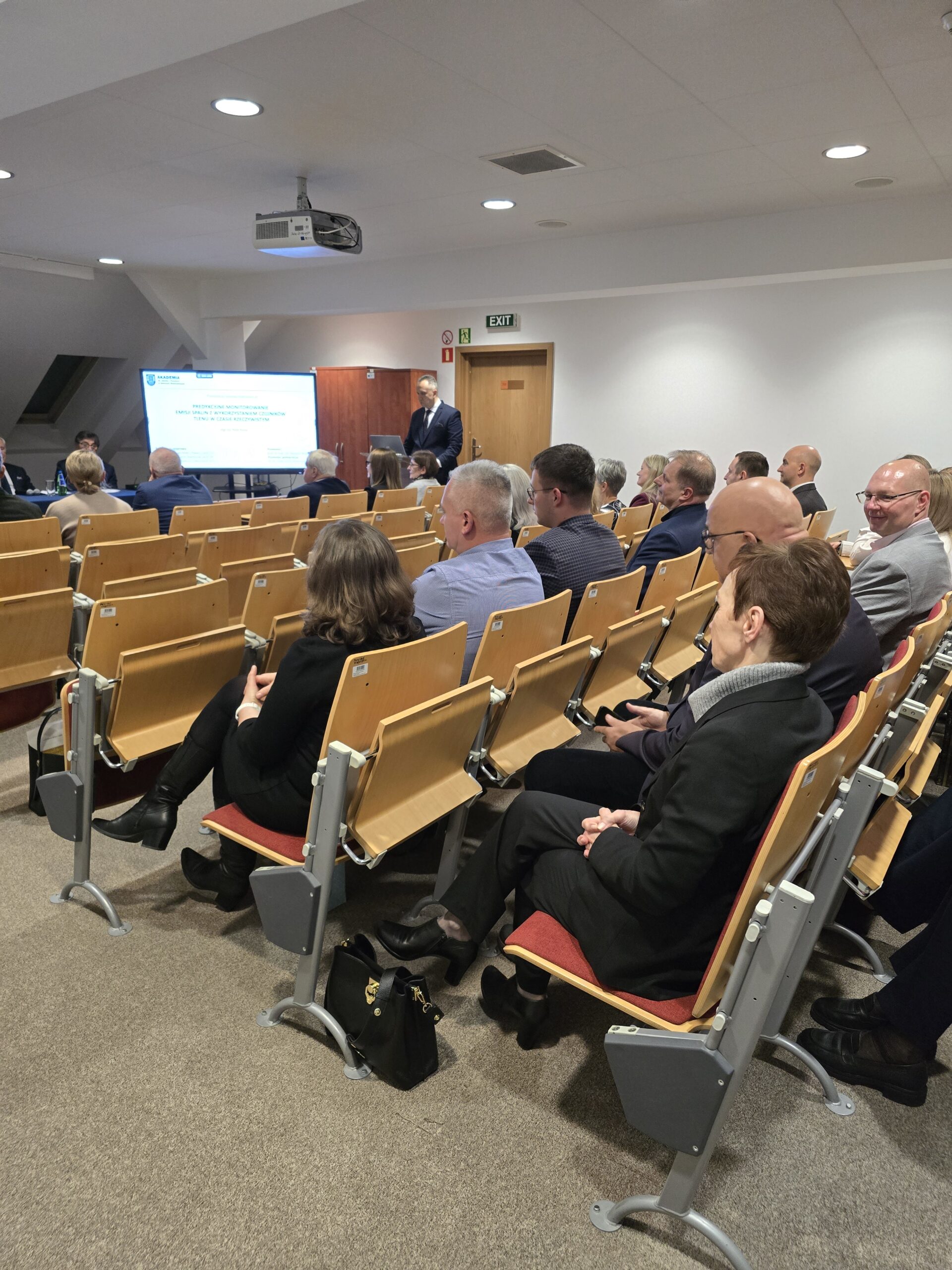 Zdjęcie sali wykładowej. Za mównicą doktorant podczas prezentacji przed komisją i zgromadzonymi gośćmi. Na pierwszym planie widać Panią Rektor siedzącą na sali.