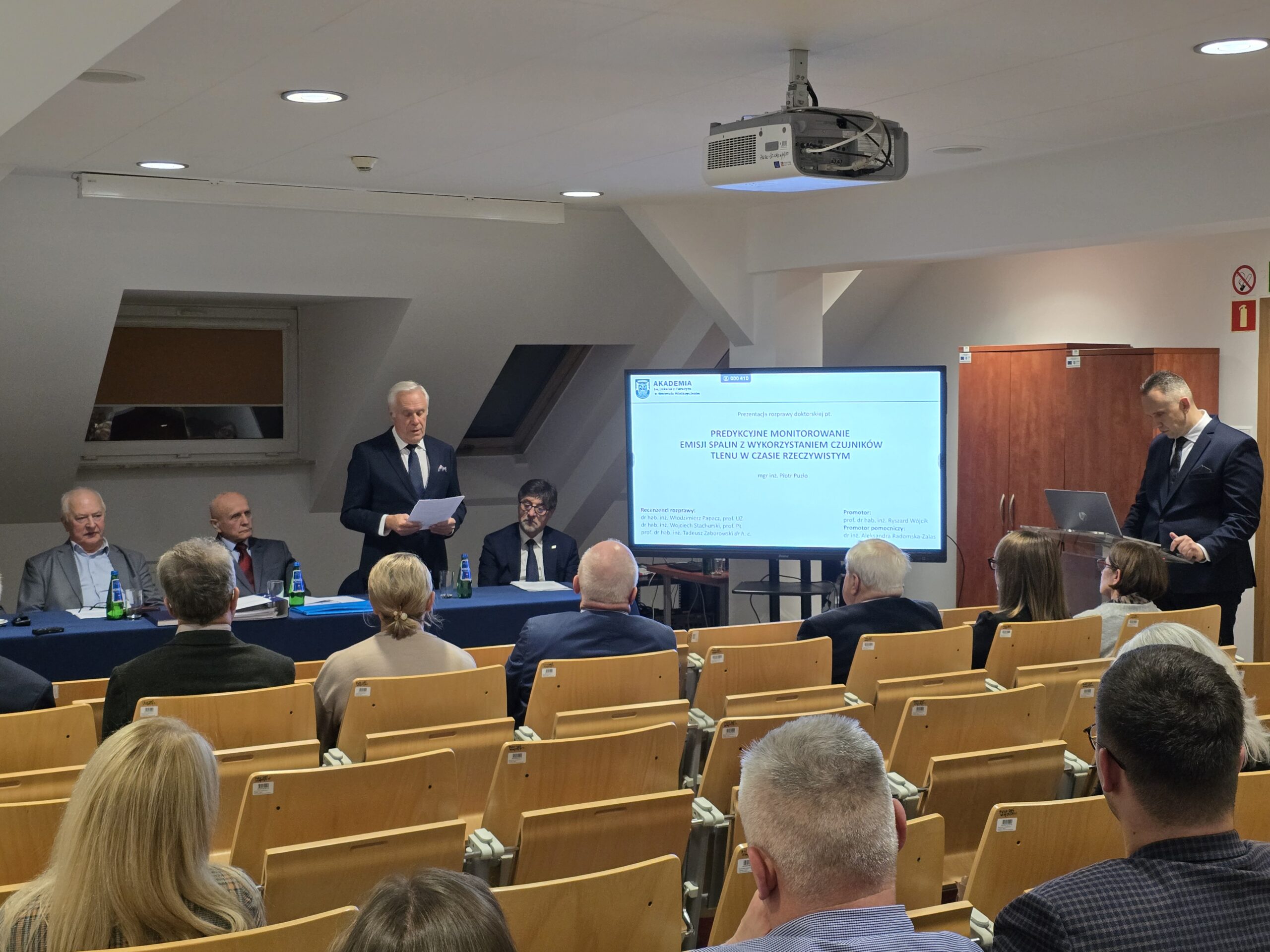 Zdjęcie sali wykładowej. Za mównicą doktorant podczas prezentacji przed komisją i zgromadzonymi gośćmi. Zabrał głos mężczyzna z komisji.