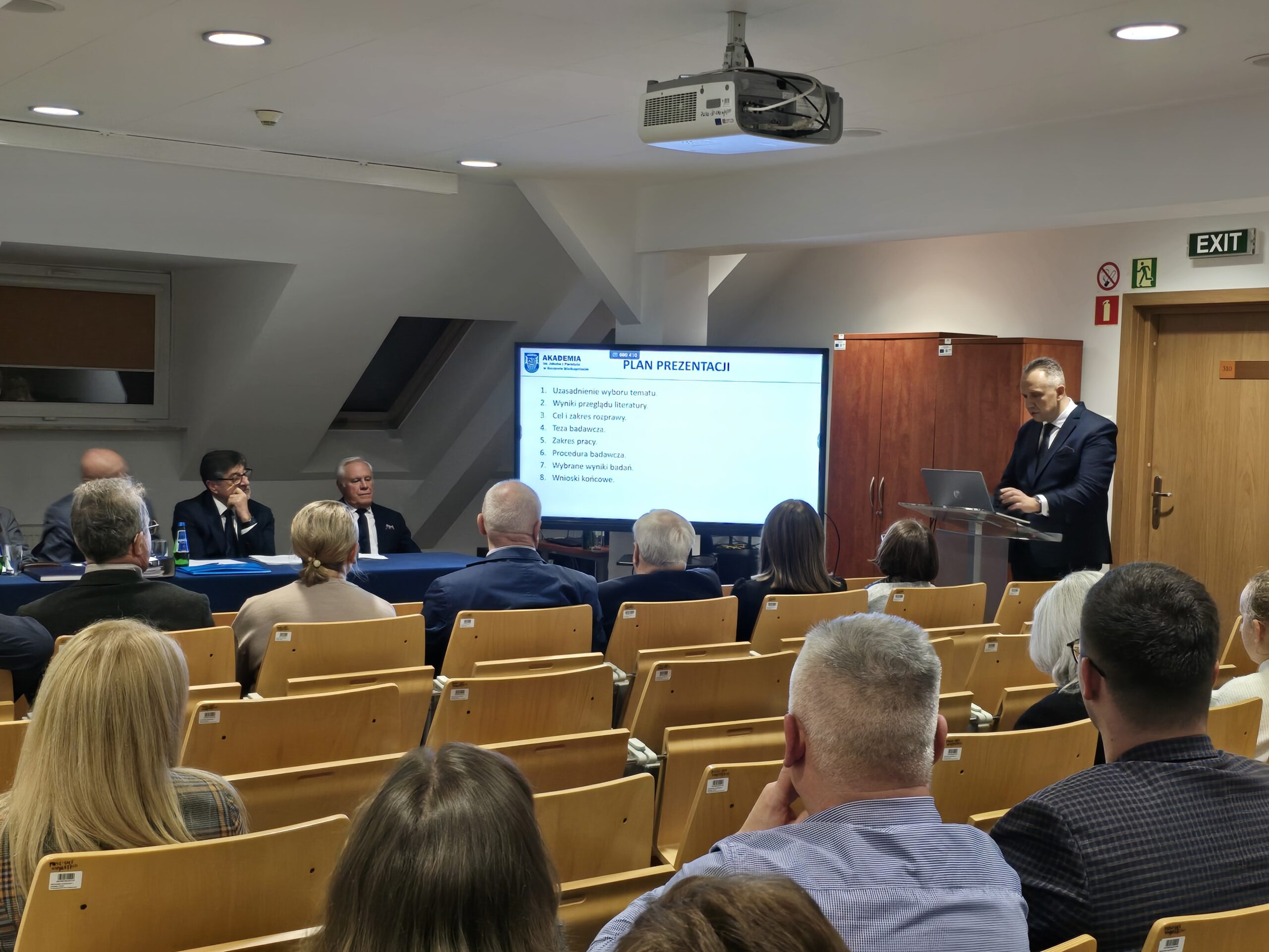 Zdjęcie sali wykładowej. Za mównicą doktorant podczas prezentacji przed komisją i zgromadzonymi gośćmi.