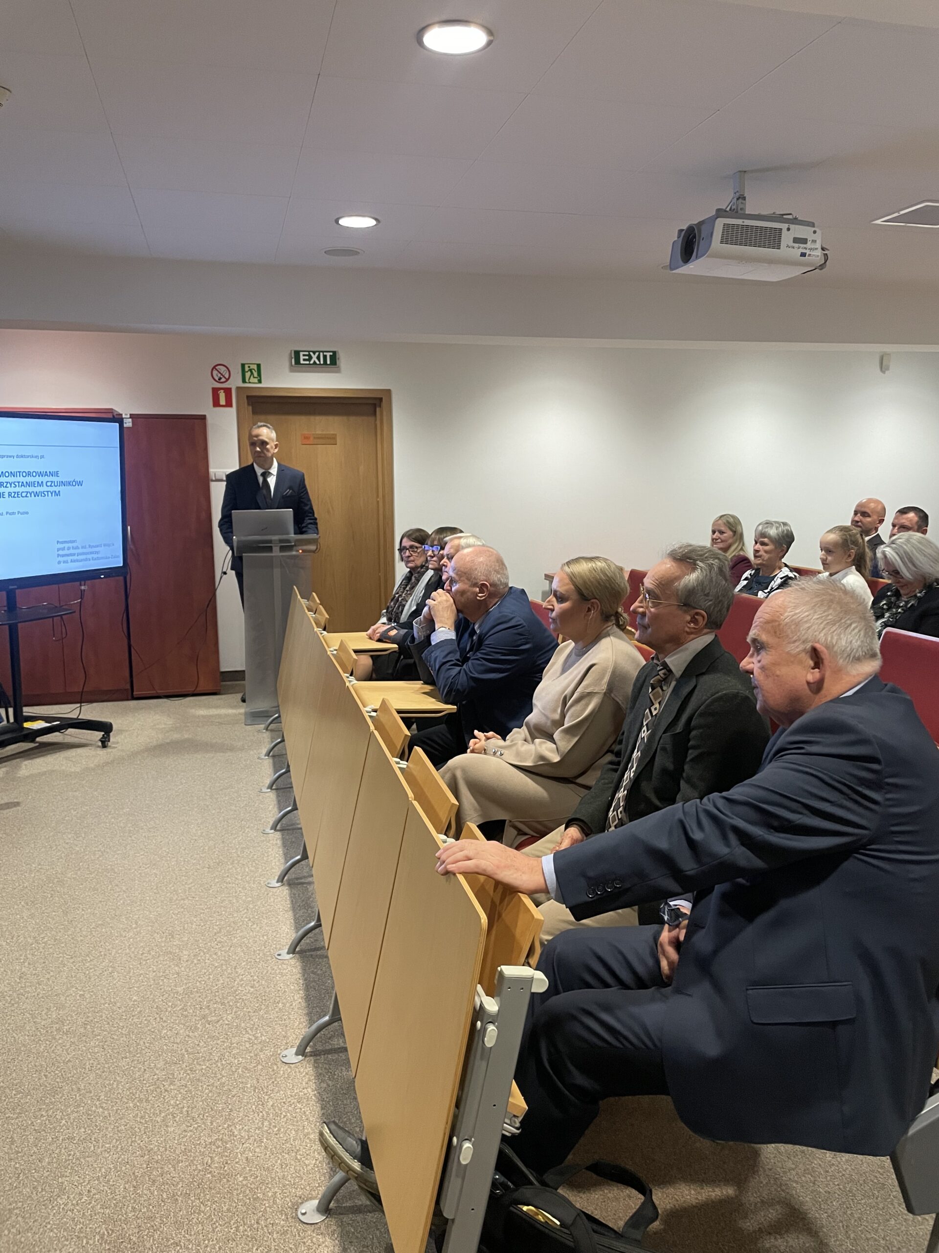 Zdjęcie sali wykładowej. Za mównicą doktorant. Na pierwszym planie widać uczestników siedzących w pierwszym rzędzie.