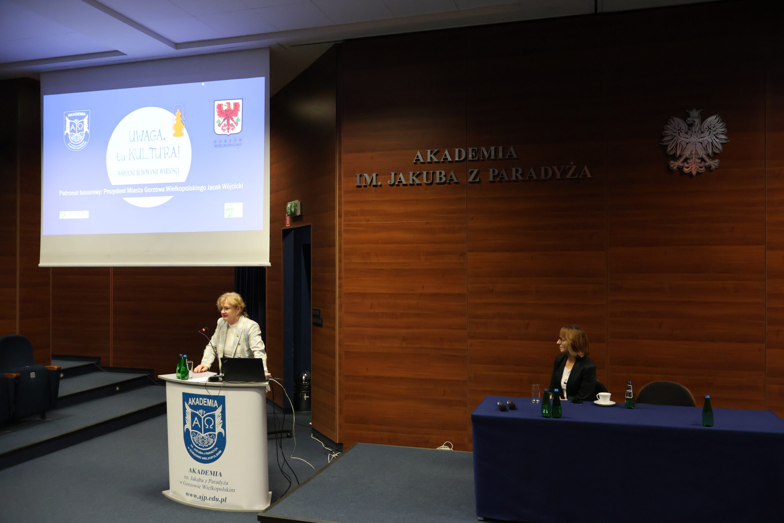 Kobieta za mównicą. Po prawej siedzi druga kobieta za stołem.