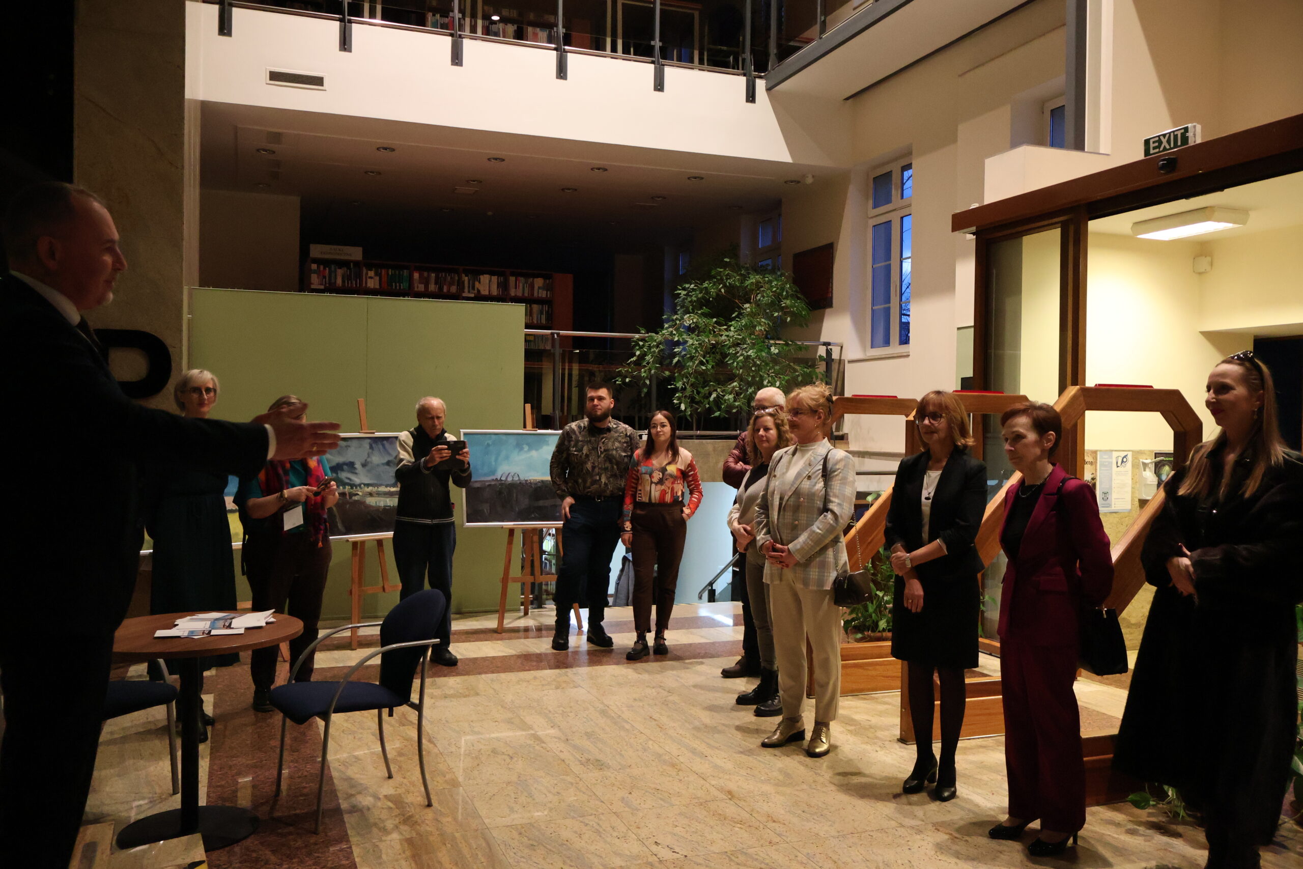 Grupa osób podczas rozpoczęcia wystawy malarstwa. Po lewej wykładowca wita przybyłych gości.
