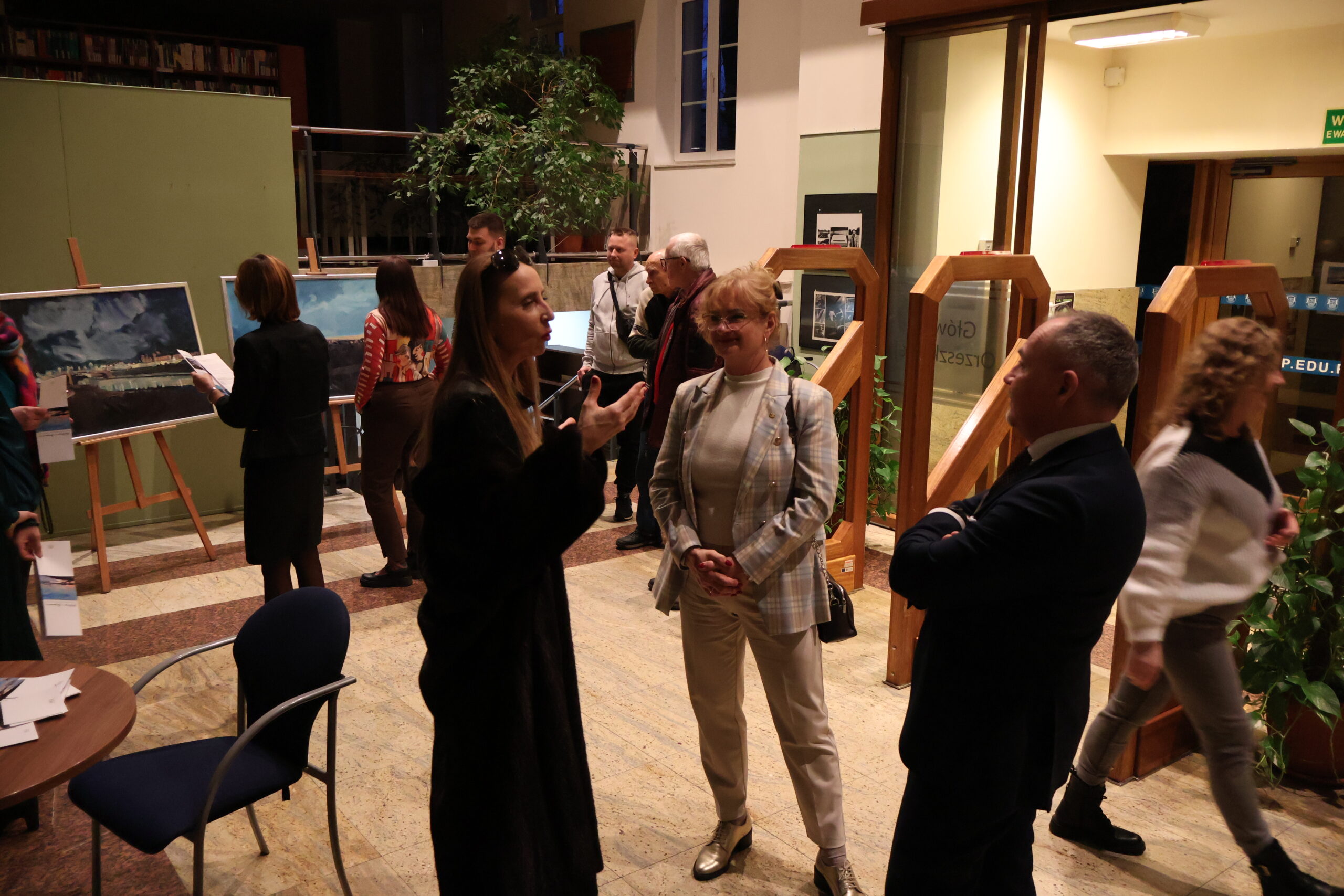 Grupa osób ogląda obrazy. Na pierwszym planie trzy osoby rozmawiają.