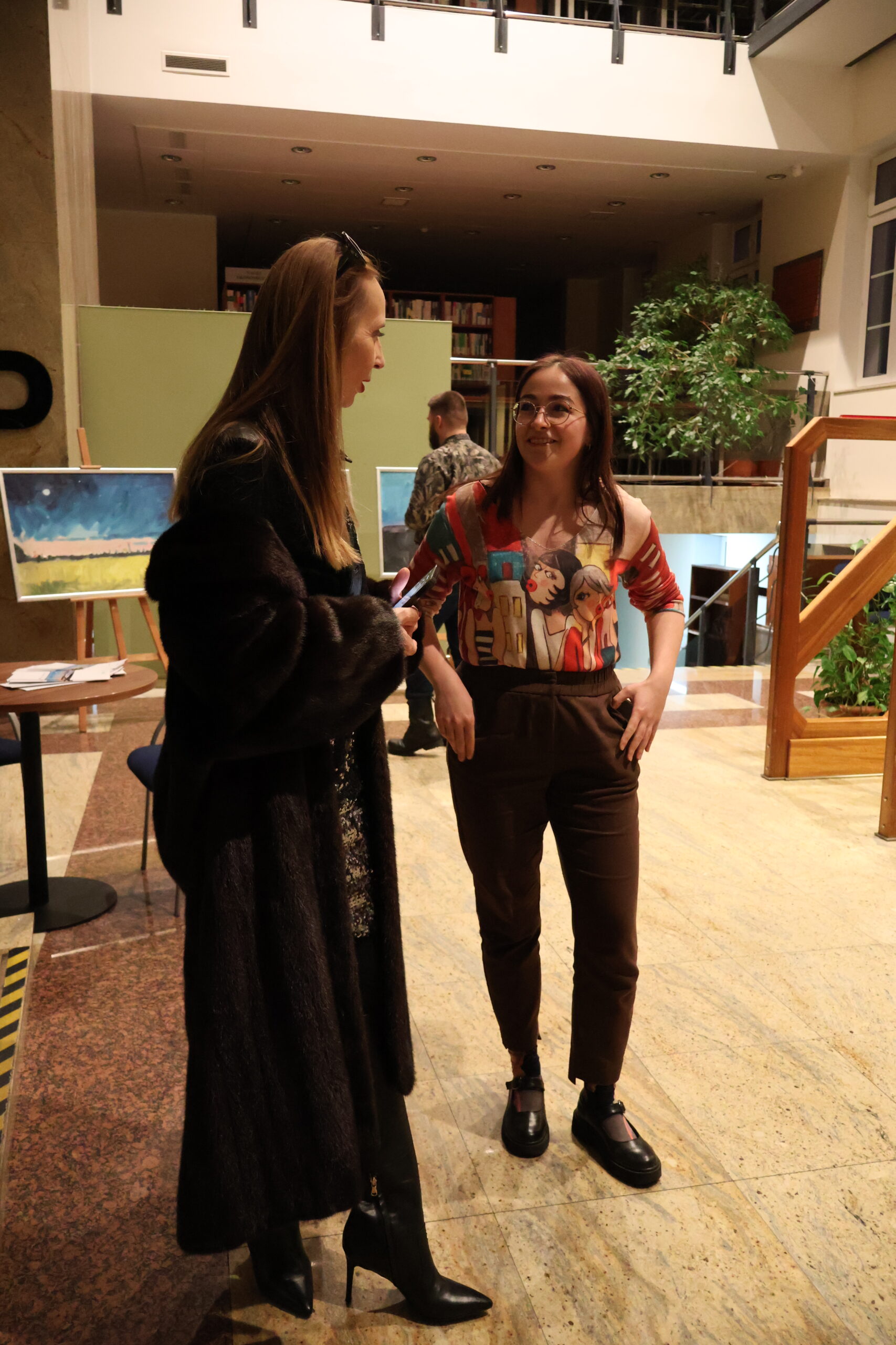 Kobieta rozmawia z malarką wystawianego wernisażu. W tle wystawa obrazów.