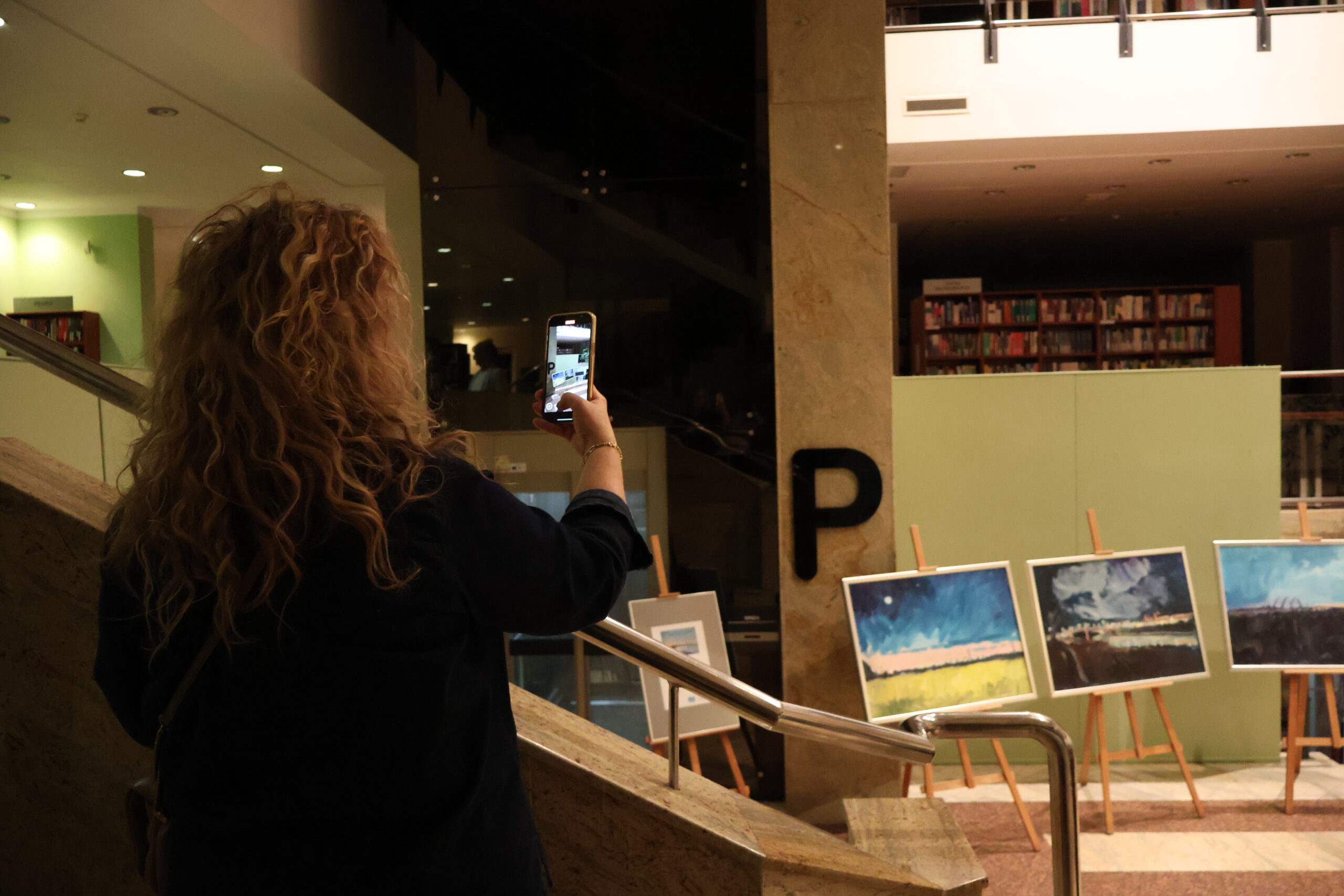 Kobieta fotografuje telefonem obrazy wystawione na wernisażu.