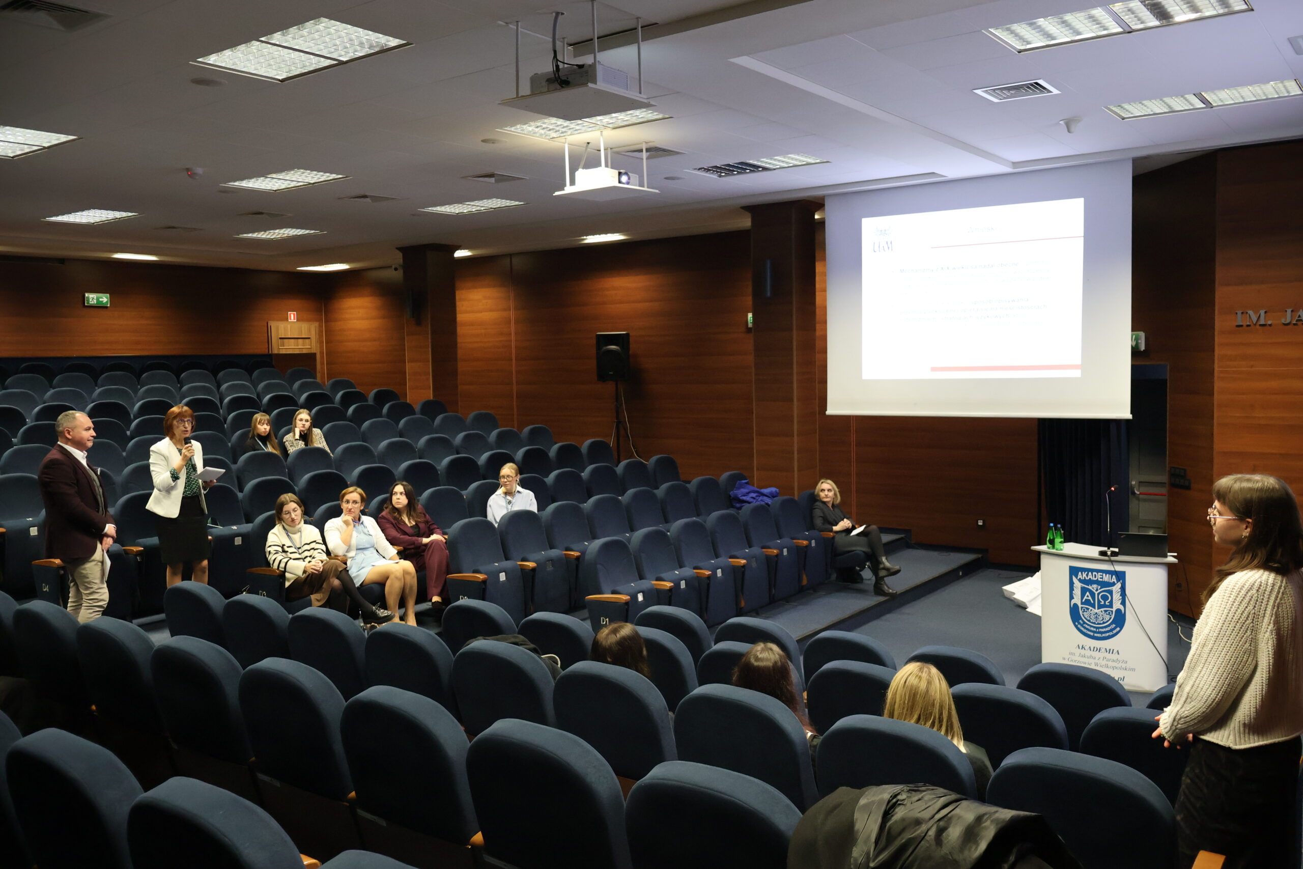 W panelu dyskusyjnym po zakończonej konferencji dr Renata Janicka-Szyszko w towarzystwie dr Wojciecha Kuski zadaje pytanie znajdującej się po drugiej stronie auli prelegentce. W tle zgromadzeni w auli goście i prelegenci.