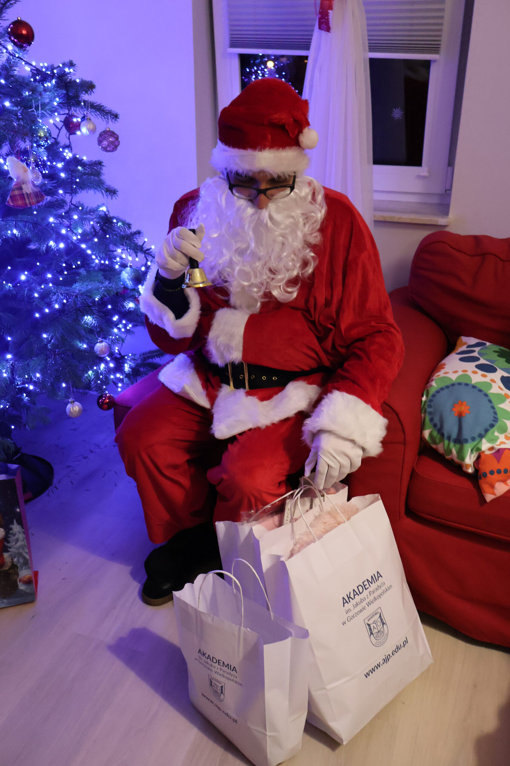 Mikołaj siedzi na pufie, w ręku trzyma dzwoneczek i patrzy na kolejne prezenty do wręczenia