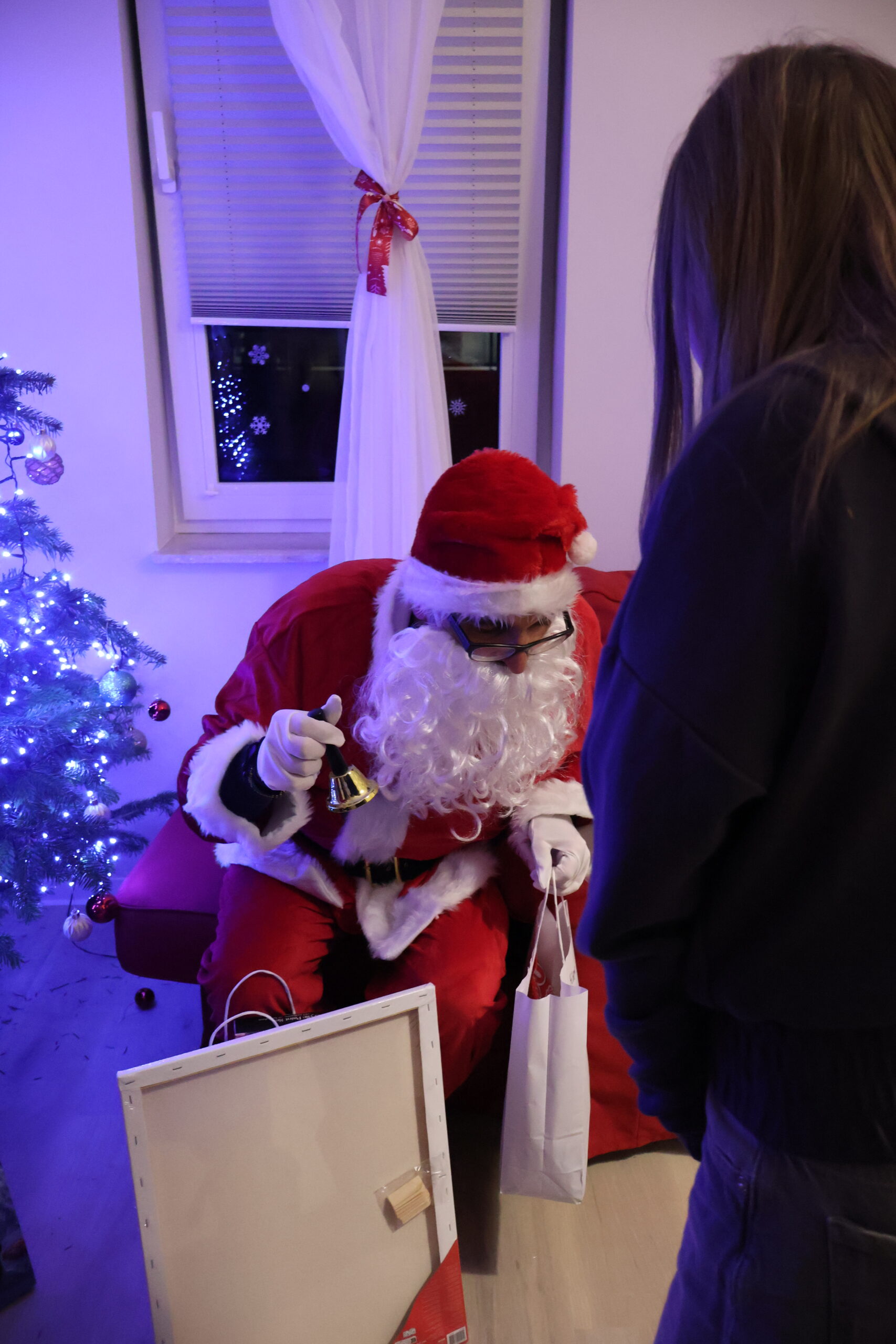 Mikołaj trzyma dzwonek w jednej ręce, a w drugiej prezent dla dziewczynki, która stoi przed nim