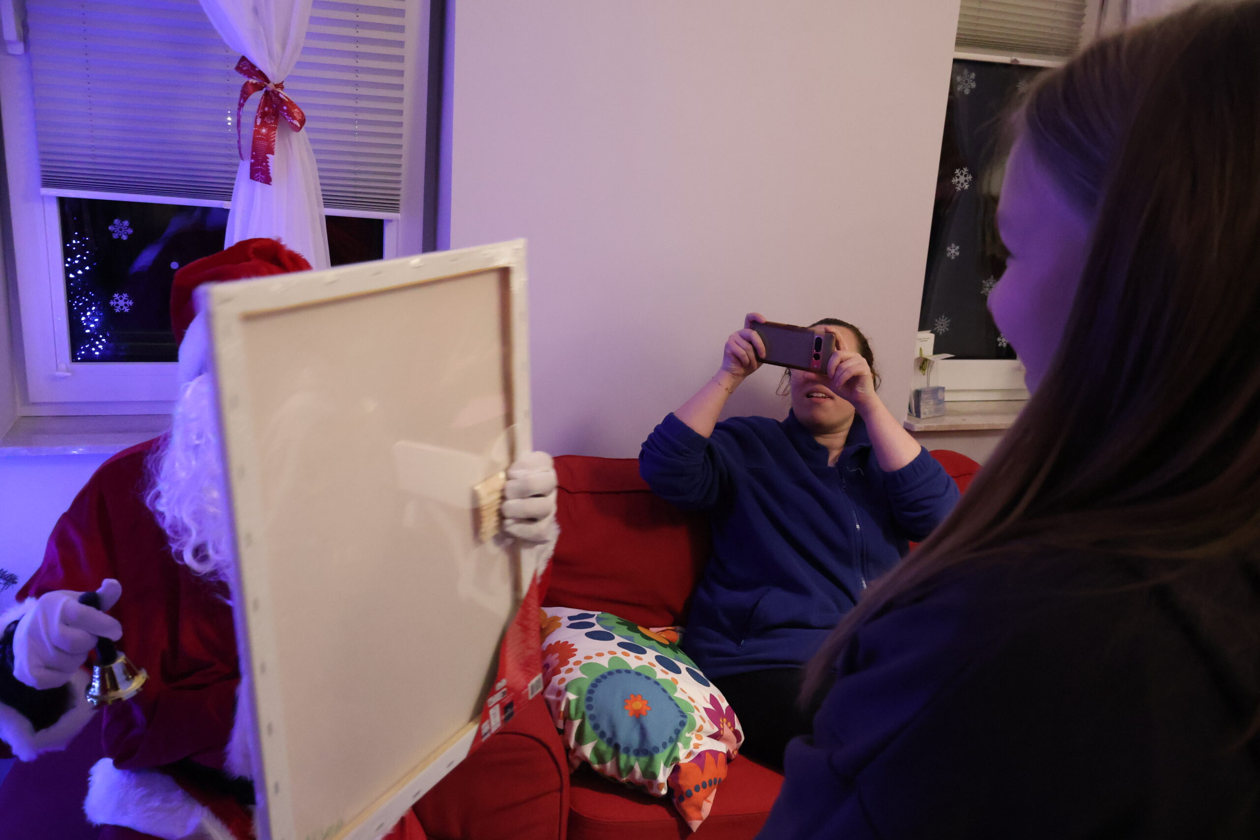 Mikołaj trzyma prezent dla stojącej przed nim dziewczynki, obok na kanapie siedzi osoba robiąca zdjęcie telefonem komórkowym