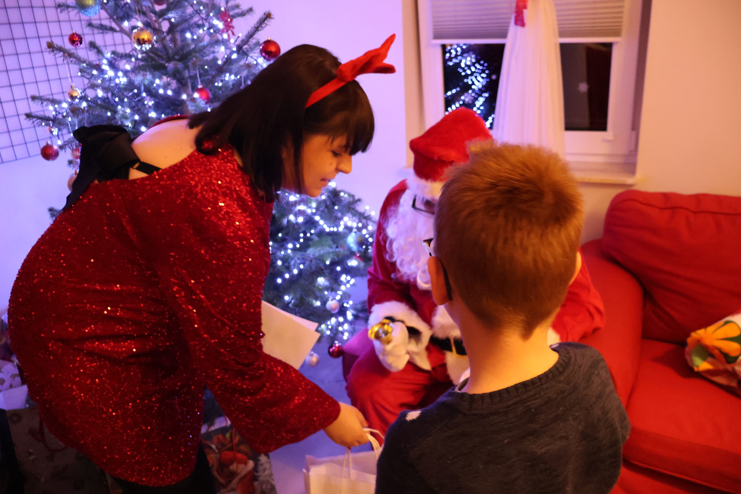 Pomocnica Mikołaja, po lewej stronie na zdjęciu, podaje prezent chłopcu, który stoi przed Mikołajem