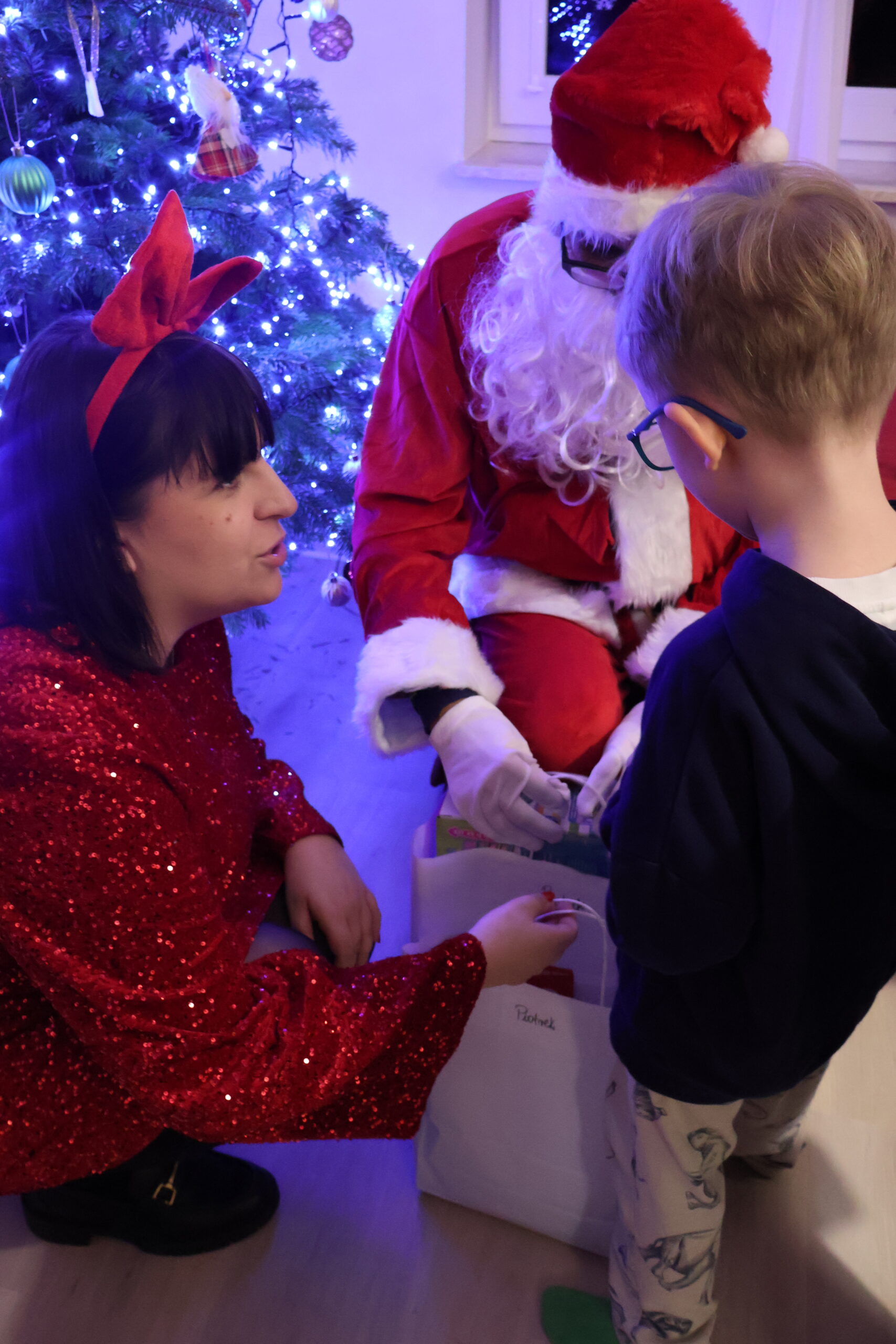 Pomocnica Mikołaja, w czerwonej sukience, kuca obok Mikołaja i dziecka, które odbiera prezenty