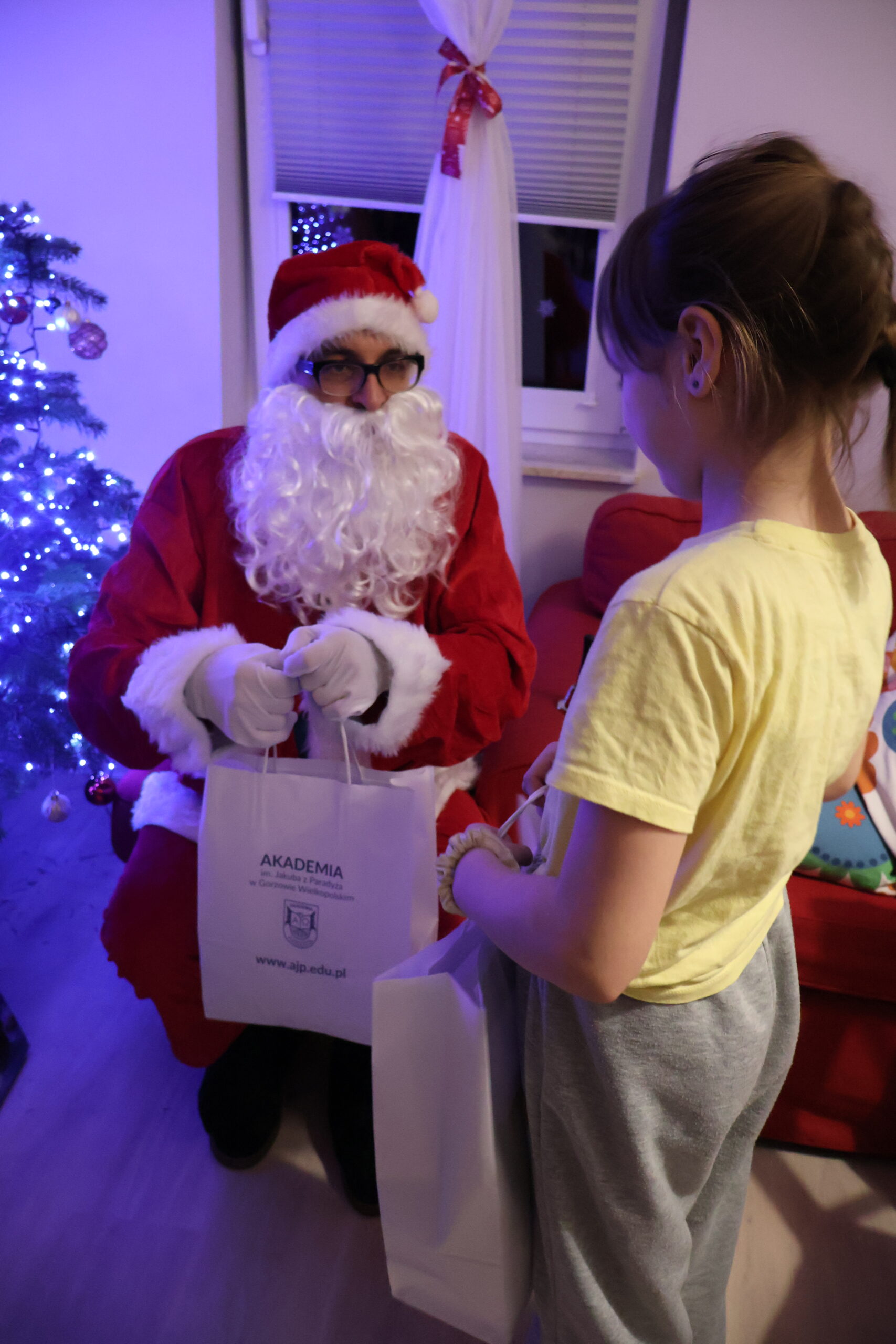 Akademicki Mikołaj siedzi na pufie i wręcza dziewczynce ze świetlicy "Jaskółka" prezent