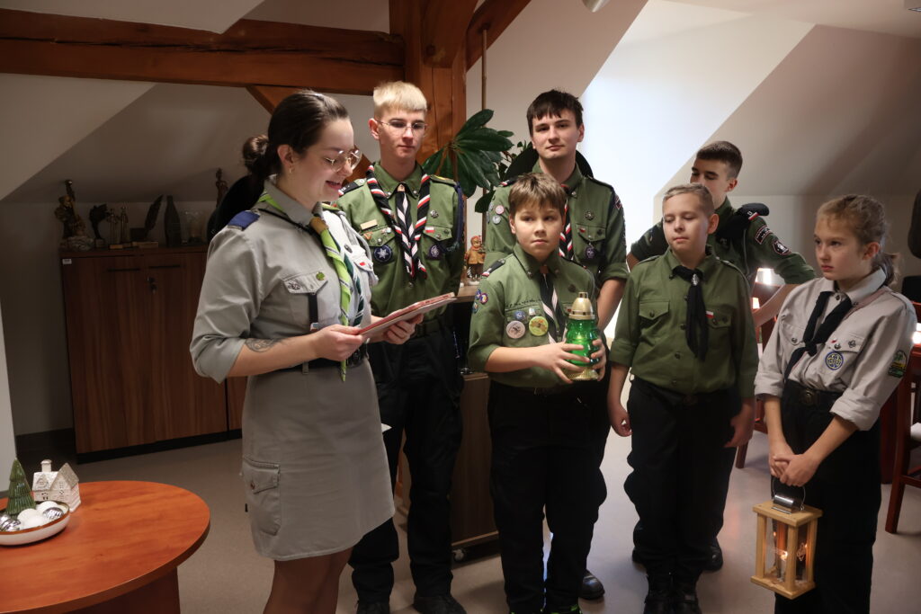 Grupa harcerzy z Gorzowa stoi w Rektoracie.Dwójka dzieci na pierwszym planie trzymają lampion i znicz w ręku. A po lewej przewodnicząca harcerzy odczytuje z kartki list. 