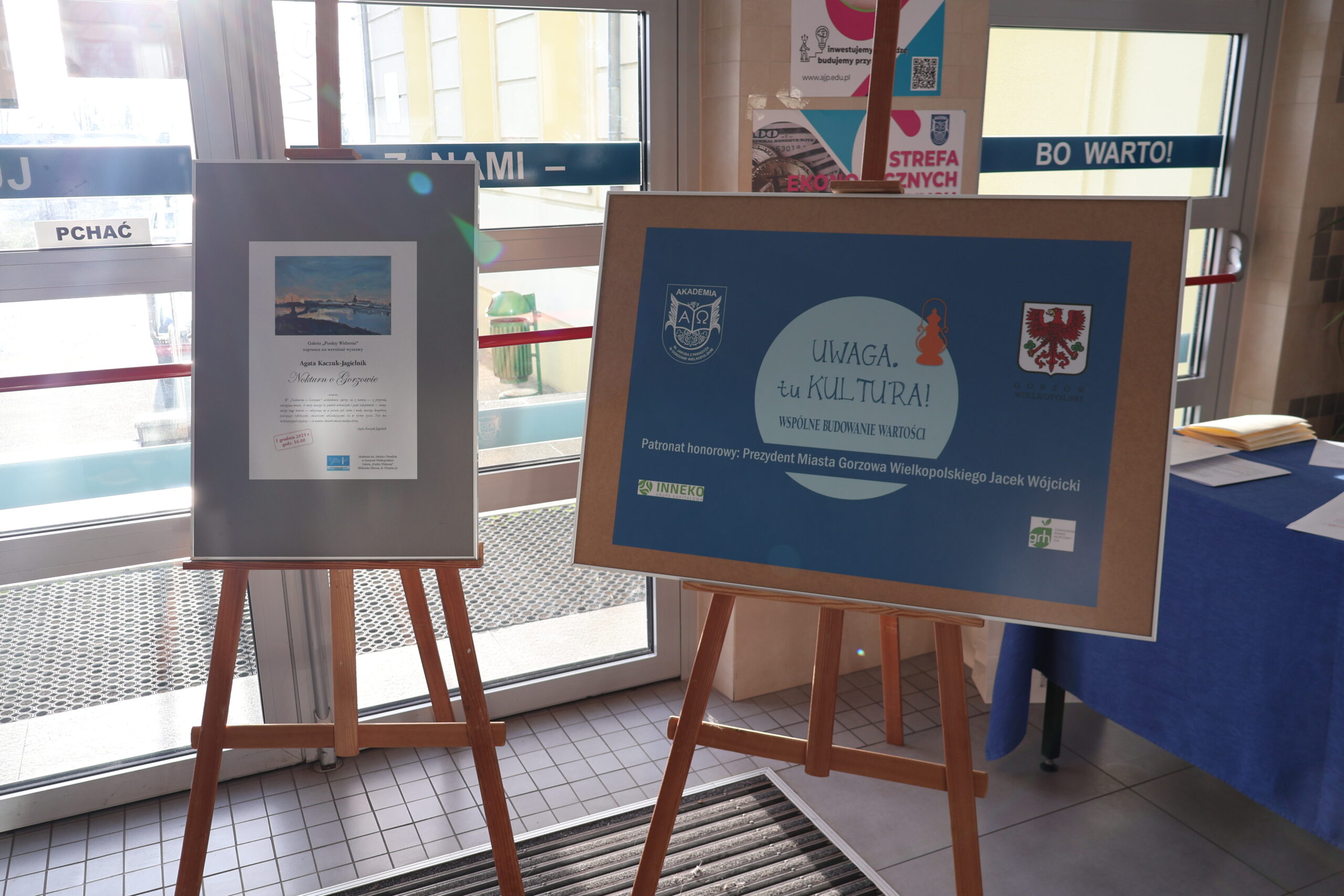 Dwa plakaty stoją na sztalugach na korytarzu. Jeden odnosi się do wystawy malarskiej, a drugi do konferencji Uwaga. tu Kultura.