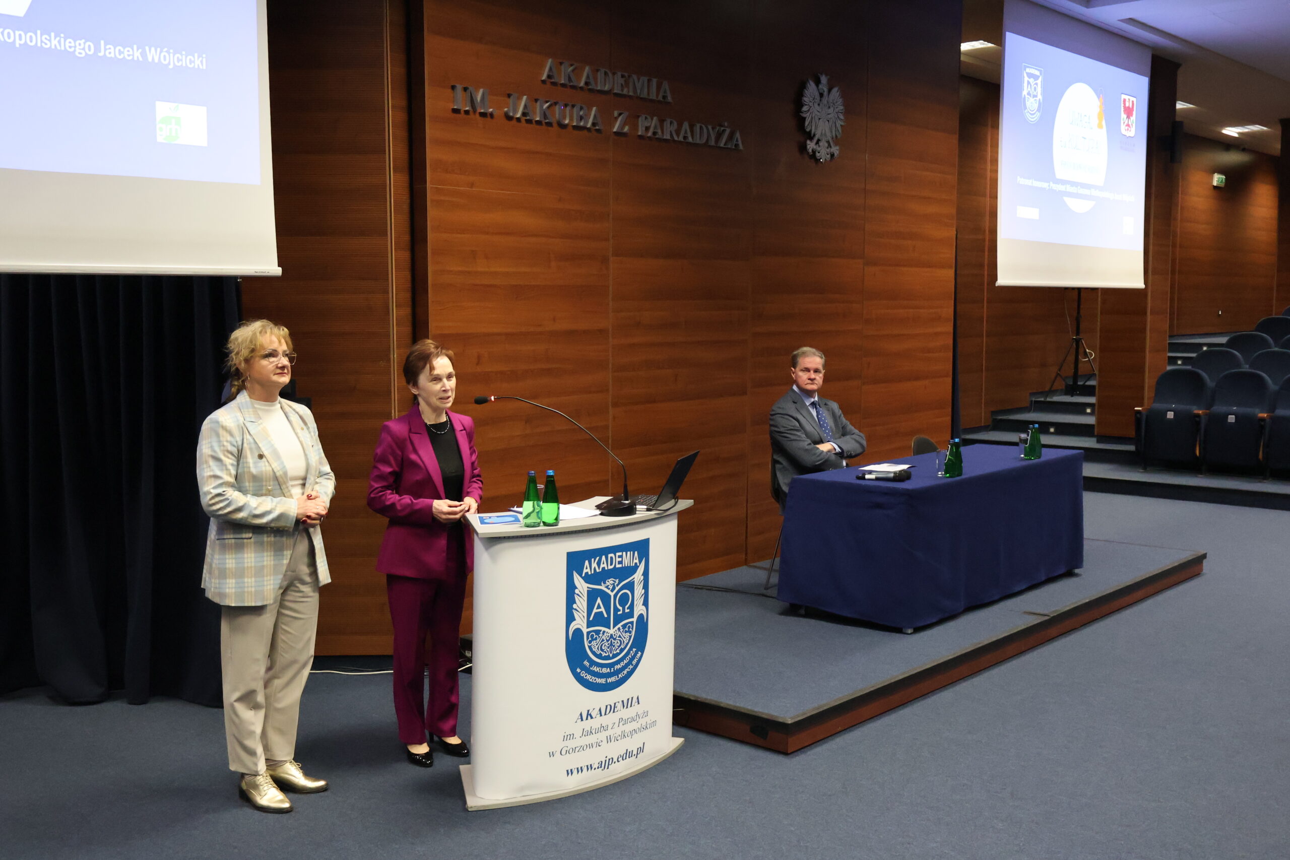 Na środku przy mównicy stoi Pani Rektor. Po lewej stoi kobieta, a po prawej za stołem siedzi Pan Prorektor.