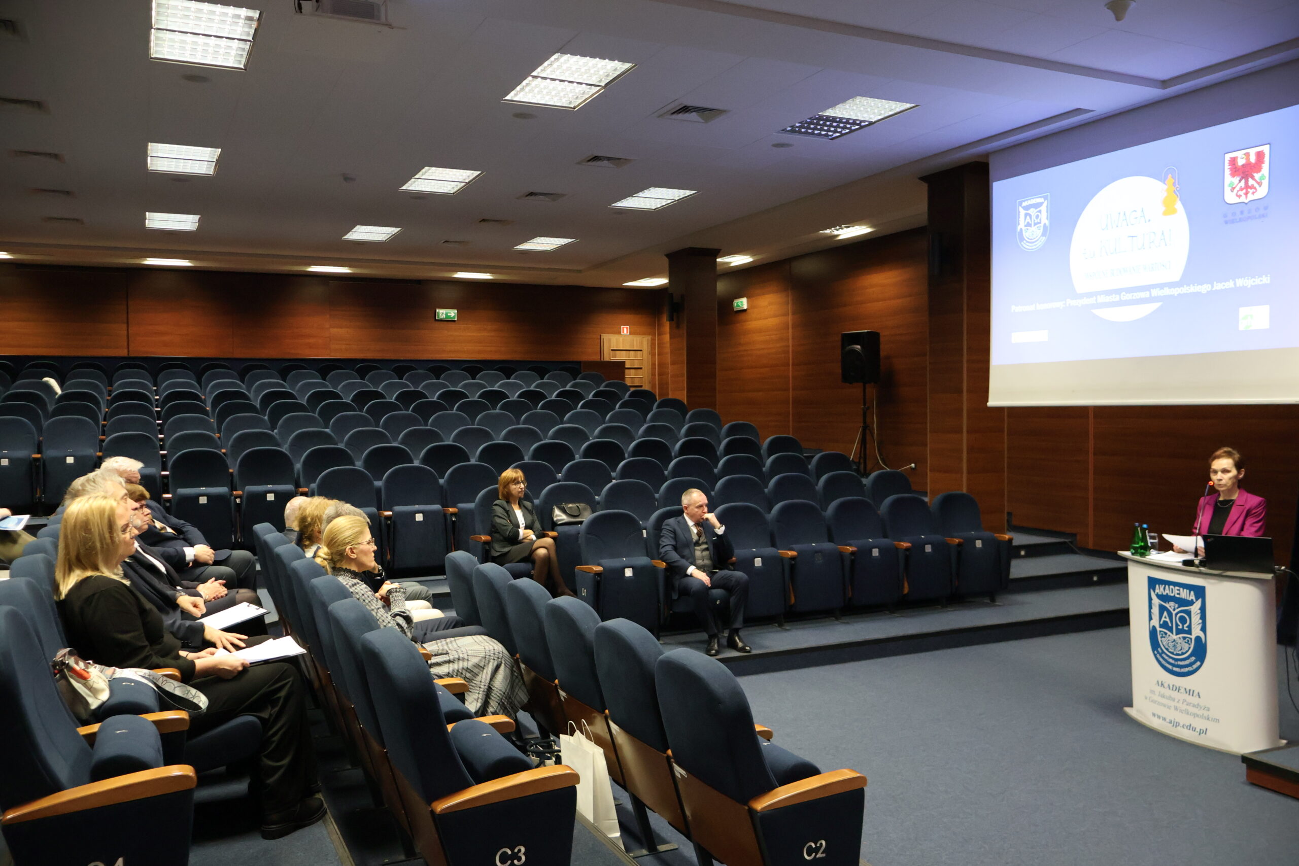 Pani Rektor stoi za mównicą podczas konferencji. Widać całą aulę z siedzącymi uczestnikami.