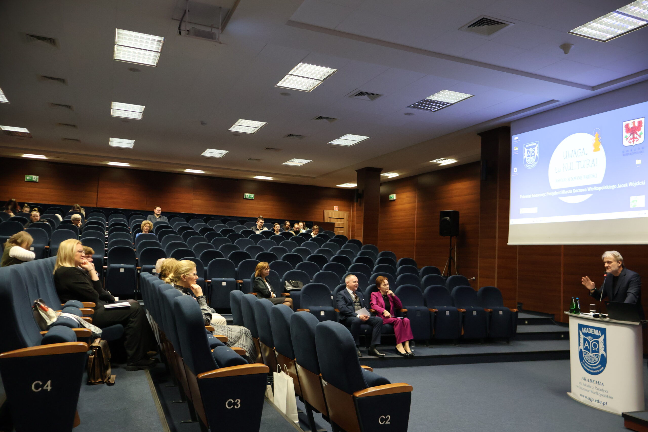 Mężczyzna stoi za mównicą podczas konferencji. Widać całą aulę z siedzącymi uczestnikami.