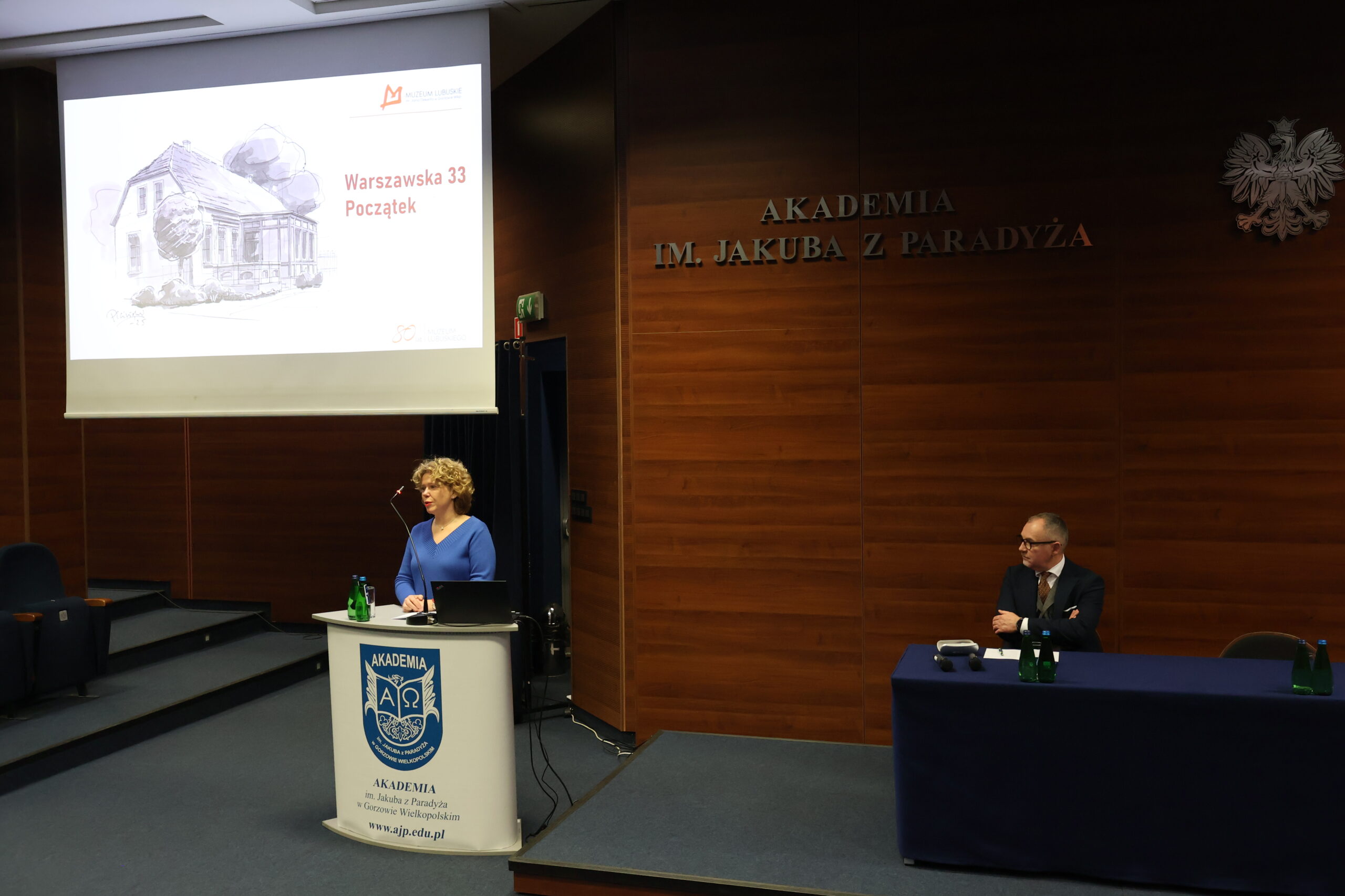 Kobieta stoi za mównicą podczas konferencji, a mężczyzna siedzi za stołem.