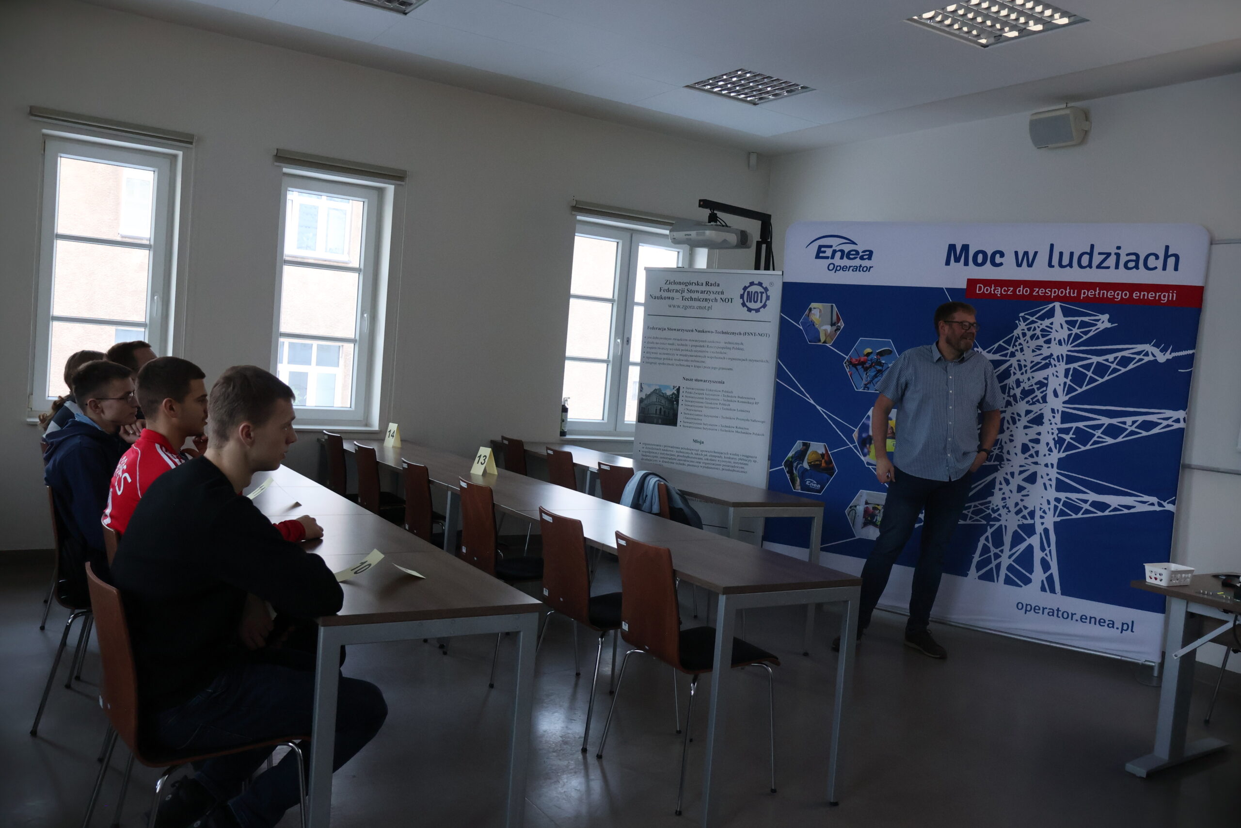 Uczestnicy olimpiady siedzą w ławkach, przed nimi stoi osoba prezentująca firmę Enea Operator.