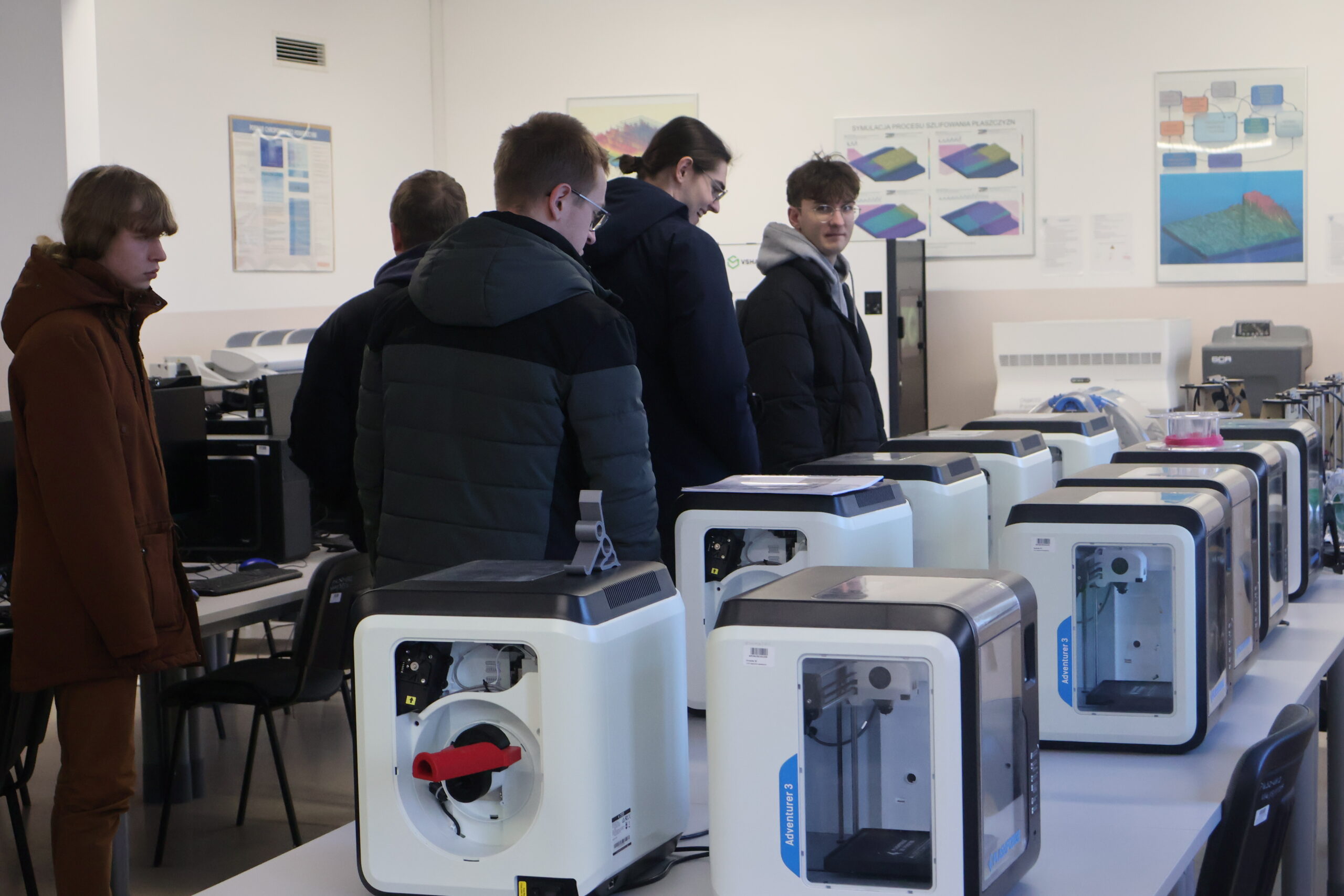 Grupa osób podczas zwiedzania laboratorium, obok nich stoją drukarki 3D.