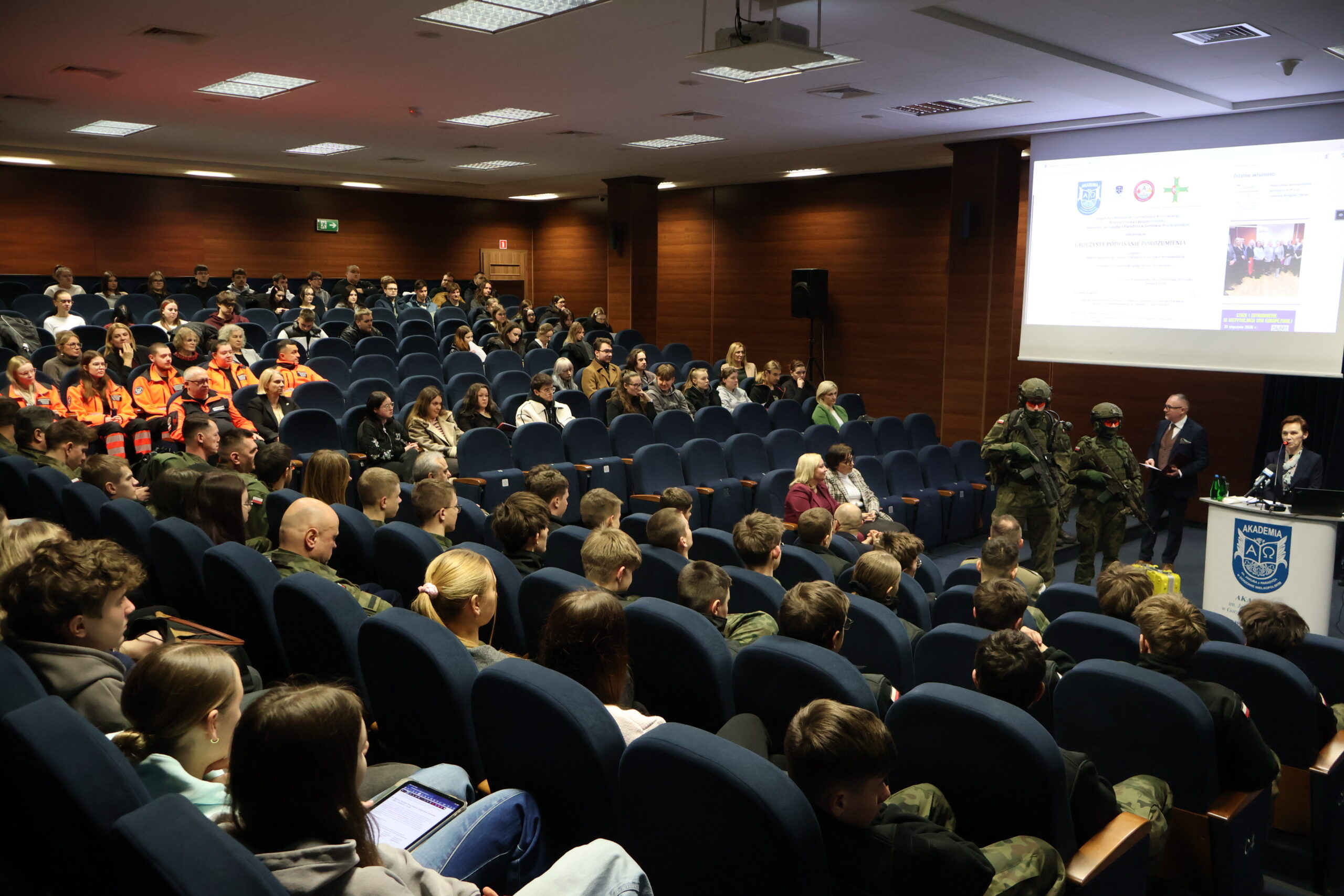 W auli im. prof. Stanisława Kirkora za mównicą wita wszystkich zgromadzonych JM Rektor prof. dr hab. Elżbieta Skorupska-Raczyńska, a obok niej dwaj żołnierze LBOT, w tle na auli zaproszeni goście, uczniowie szkół oraz pracownicy AJP