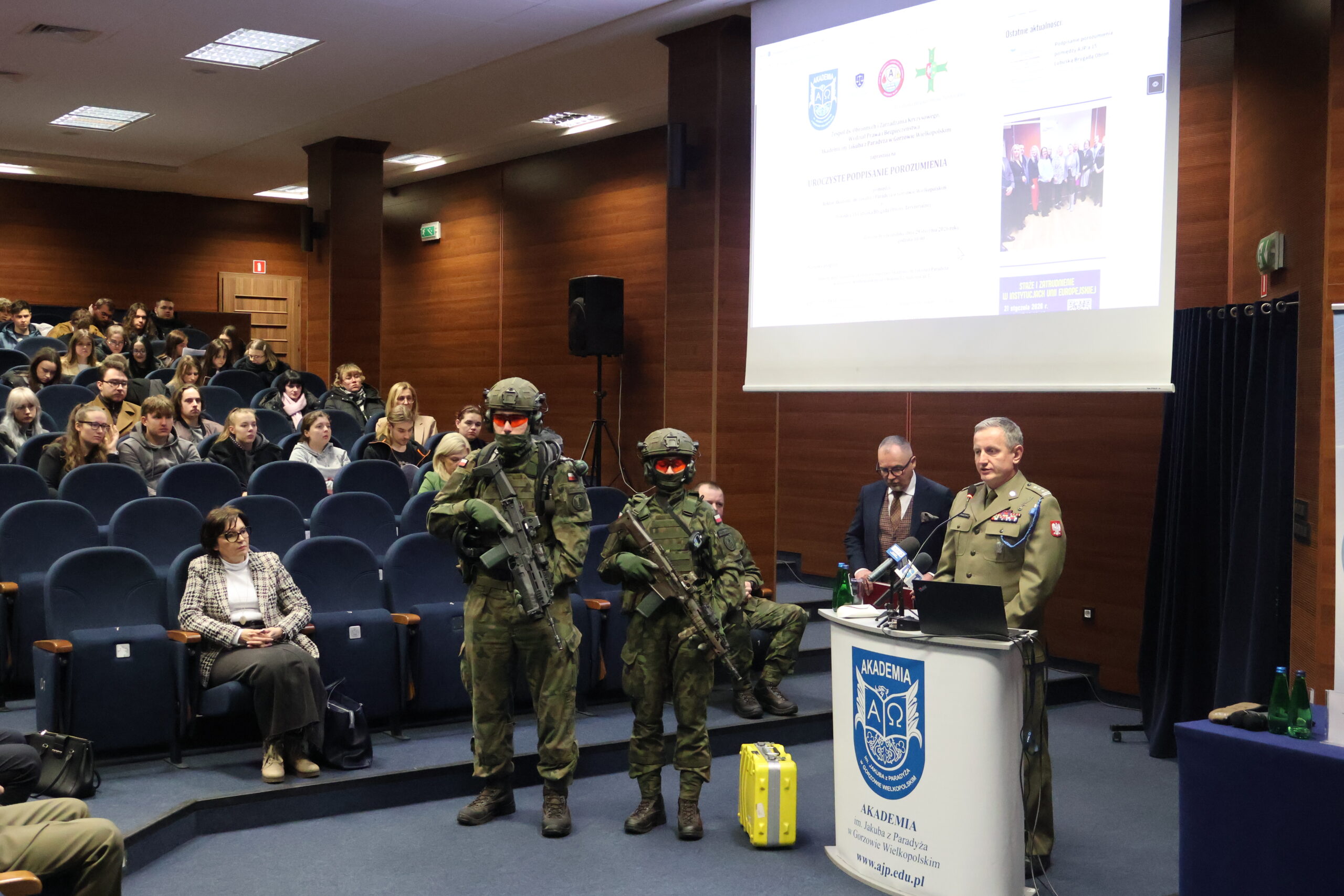 W auli im. prof. Stanisława Kirkora za mównicą stoi i przemawia w stroju służbowym płk Leszek Giergielewicz, Dowódca 15 Lubuskiej Brygady Obrony Terytorialnej, a obok niego dwaj żołnierze LBOT, w tle na auli zaproszeni goście.