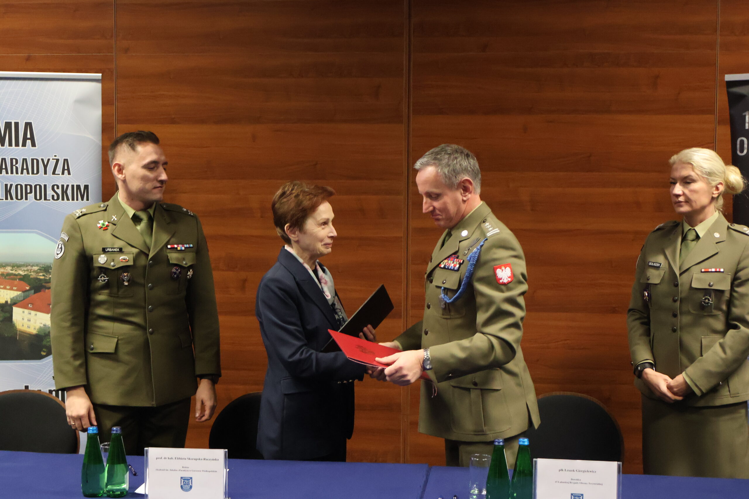 Płk Leszek Giergielewicz i prof. dr hab. Elżbieta Skorupska-Raczyńska wymieniają się dokumentami podpisania porozumienia w towarzystwie obecnych: mjr rez. mgr dypl. Roberta Urbanka i majora Magdaleny Goliszek.
