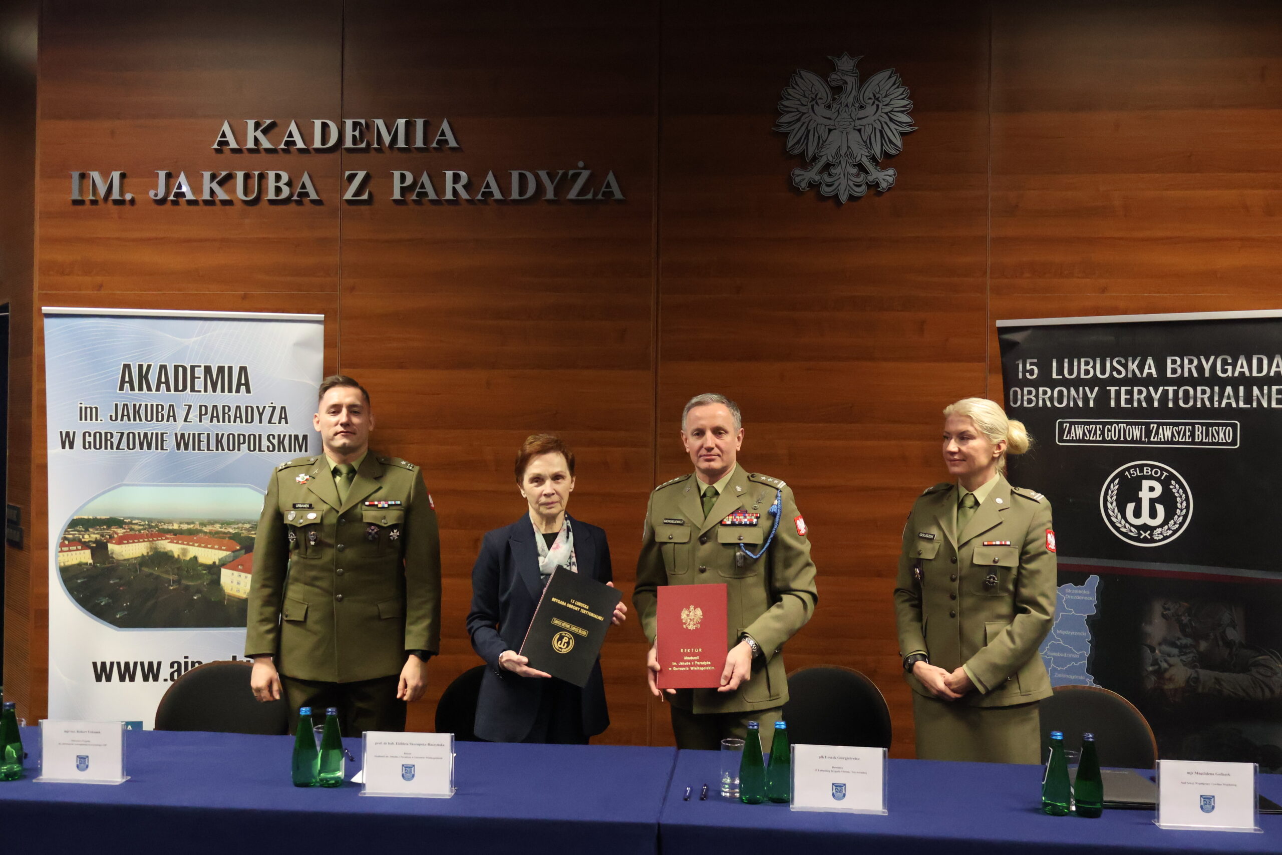 W auli im. prof. Stanisława Kirkora za stołem prezydialnym - prof. dr hab. Elżbieta Skorupska-Raczyńska wraz z obecnym po prawej po prawej płk Leszkiem Giergielewiczem trzymajacy dokument podpisania porozumienia, po lewej od Pani Rektor: mjr rez. mgr dypl. Robert Urbanek, a po prawej od płk stoi major Magdalena Goliszek.