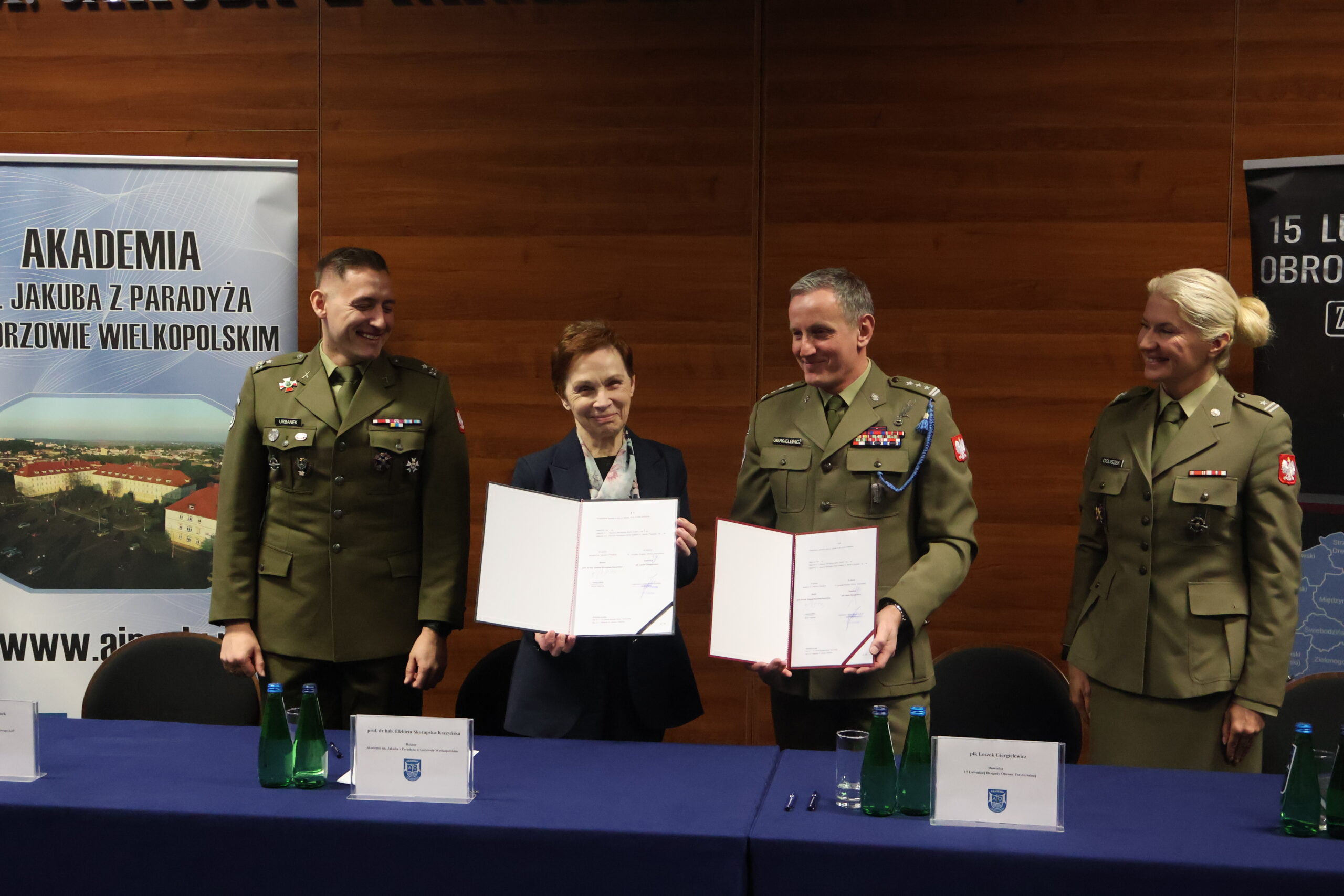 W auli im. prof. Stanisława Kirkora za stołem prezydialnym - prof. dr hab. Elżbieta Skorupska-Raczyńska wraz z obecnym po prawej po prawej płk Leszkiem Giergielewiczem trzymajacy otwarty dokument podpisania porozumienia, po lewej od Pani Rektor: mjr rez. mgr dypl. Robert Urbanek, a po prawej od płk stoi major Magdalena Goliszek.