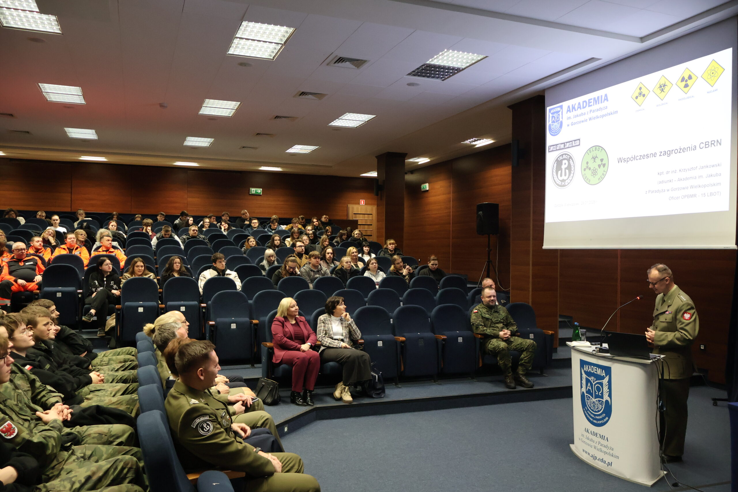 Na zdjęciu kpt. dr inż. Krzysztof Jankowski – oficer OPBMR (CBRN) w 15 Lubuskiej Brygadzie Obrony Terytorialnej; Dyrektor Instytutu Nanotechnologii i Nanobiologii AJP w stroju służbowum podczas swojego wykładu w ramach podpisania porozumienia, stoi za mównicą prezentując swój wykład pod nazwą "Współczesne zagrożenia CBRN" w auli im. prof. Stanisława Kirkora. W tle na auli w rzędach zaproszeni goście.