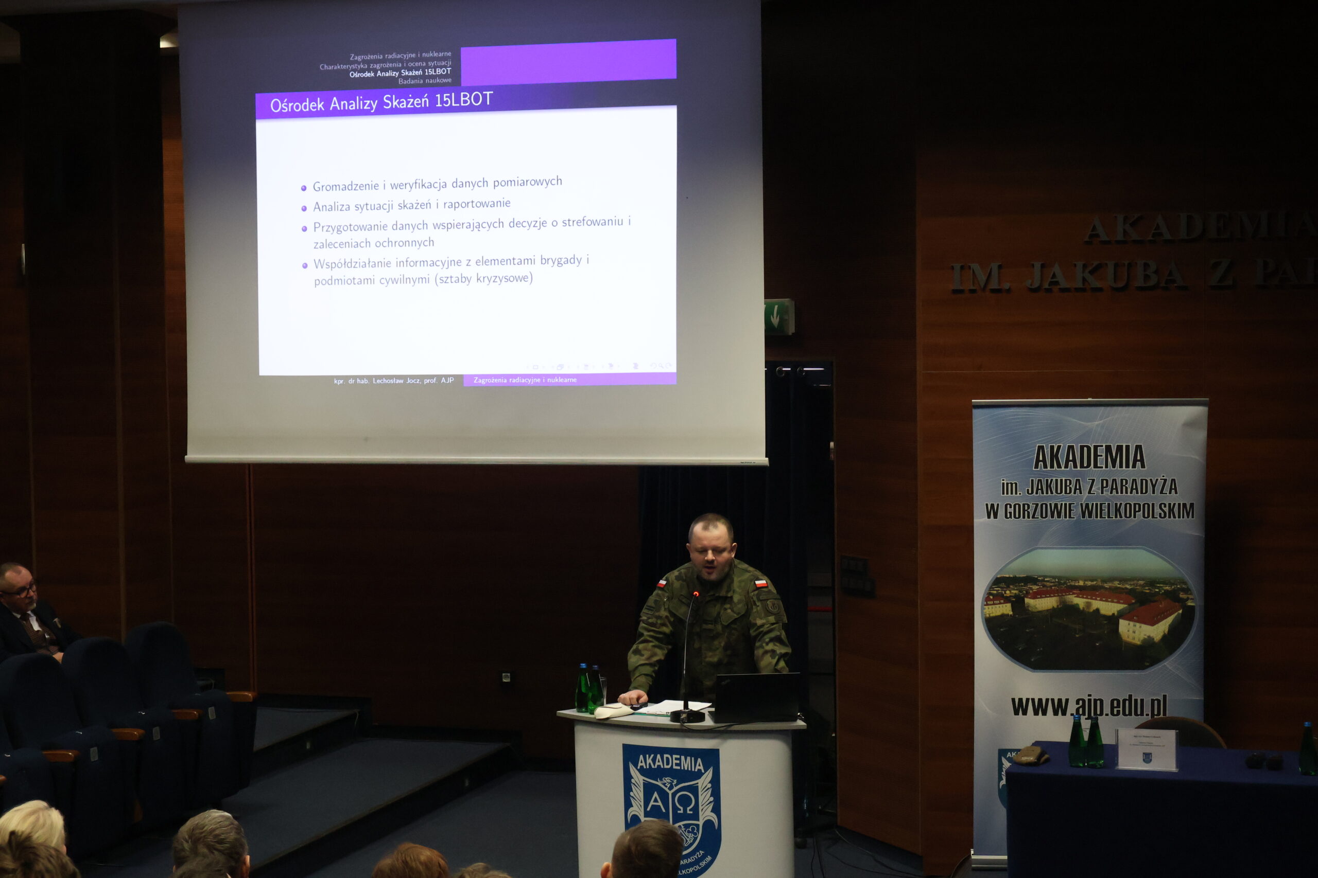 Na zdjęciu kpr. dr hab. Lechosław Jocz, prof. uczelni – analityk Ośrodka Analizy Skażeń w 15 Lubuskiej Brygadzie Obrony Terytorialnej; profesor uczelni (AJP) prezentujący wykład w ramach uroczystości podpisania porozumienia. W tle slajd z napisem Ośrodek Analizy Skażeń 15 LBOT.