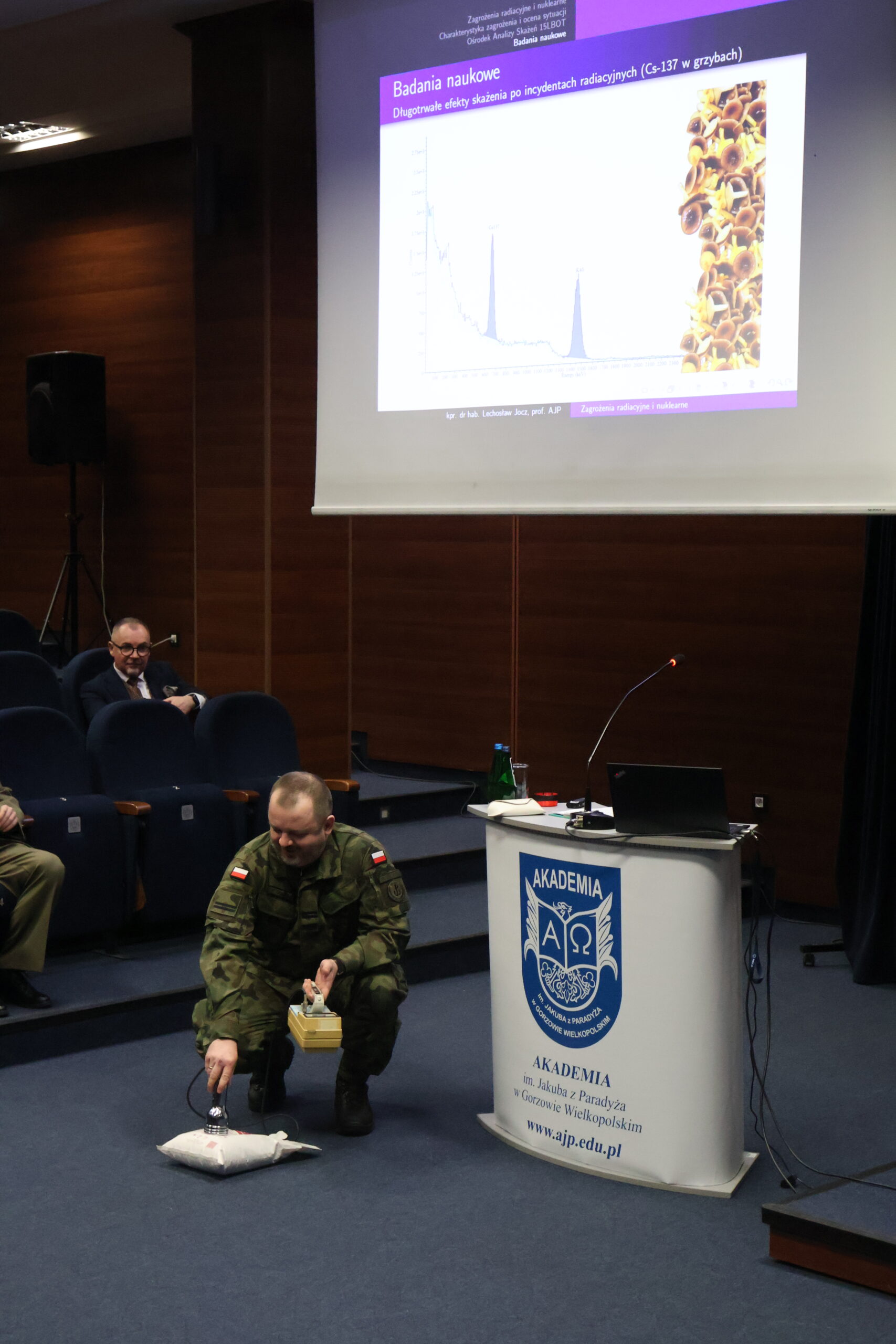 Na zdjęciu kpr. dr hab. Lechosław Jocz, prof. uczelni – analityk Ośrodka Analizy Skażeń w 15 Lubuskiej Brygadzie Obrony Terytorialnej; profesor uczelni (AJP) prezentujący wykład w ramach uroczystości podpisania porozumienia. W tle slajd z napisem Badania naukowe. Kapral klęka ze sprzętem i pokazuje jaki wydobywa się dźwięk.