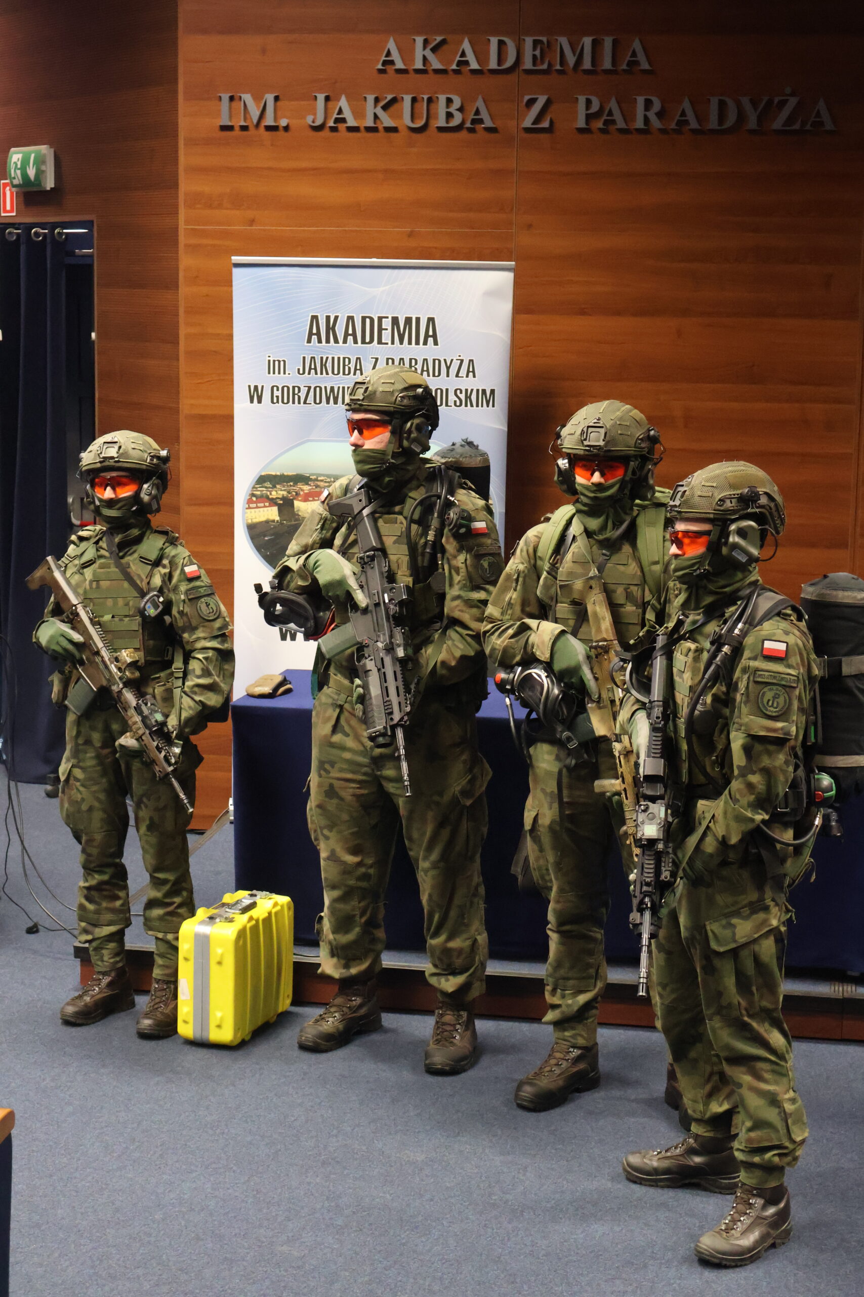 Na zdjęciu widoczni są czterej żołnierze z 15 LOBT ubrani w mundury wojskowe z karabinami w auli im. prof. Stanisława Kirkora. Na tle napis Akademia im. Jakuba z Paradyża na ścianie i na roll-upie.