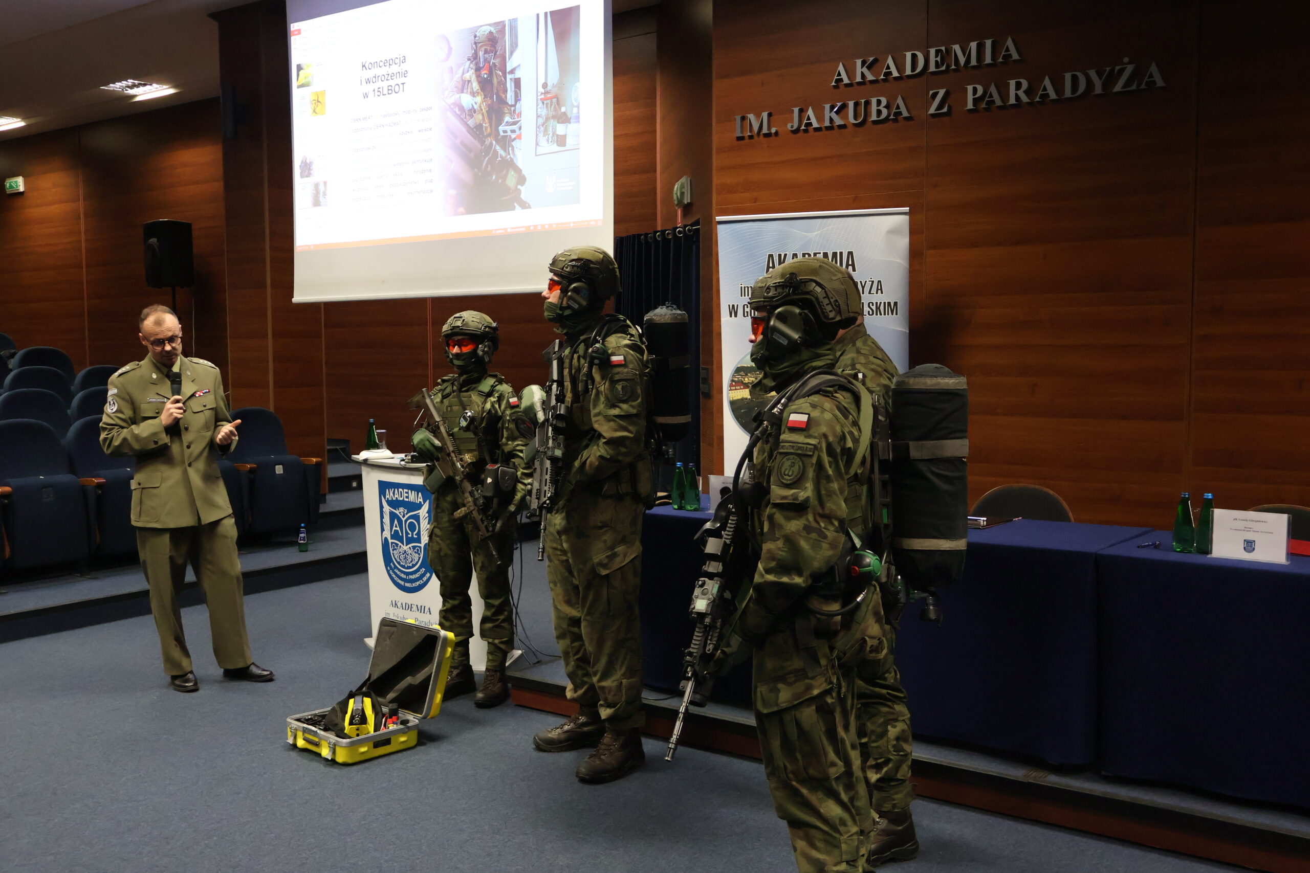 Na zdjęciu kpt. dr inż. Krzysztof Jankowski – oficer OPBMR (CBRN) w 15 Lubuskiej Brygadzie Obrony Terytorialnej; Dyrektor Instytutu Nanotechnologii i Nanobiologii AJP w stroju służbowum podczas swojego wykładu w ramach podpisania porozumienia, stoi z mikrofonem przy 4 żołnierzach z 15 LOBT ubranych w mundury wojskowe z karabinami w auli im. prof. Stanisława Kirkora.