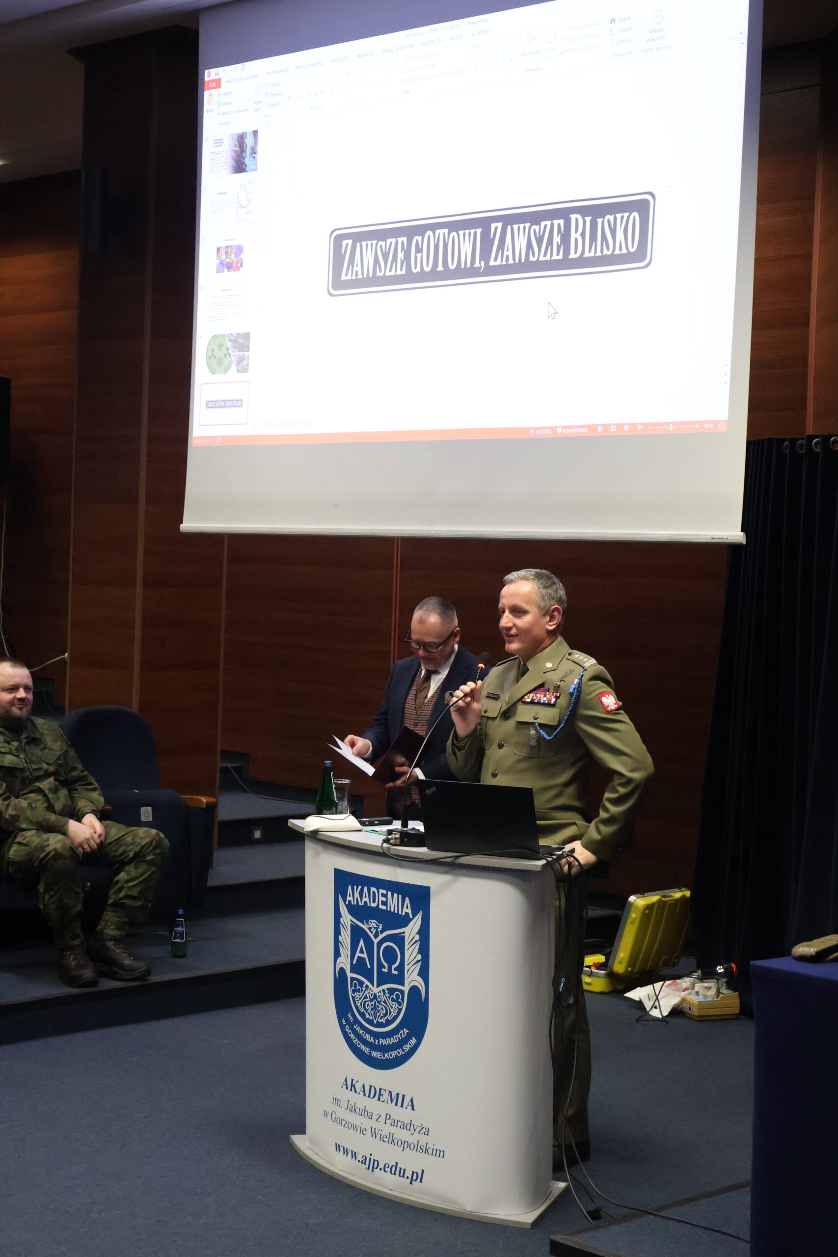 W auli im. prof. Stanisława Kirkora za mównicą stoi i przemawia na zakończenie w stroju służbowym płk Leszek Giergielewicz, Dowódca 15 Lubuskiej Brygady Obrony Terytorialnej, a za nim slajd z napisem"ZAWSZE GOTOWI,ZAWSZE BLISKO"