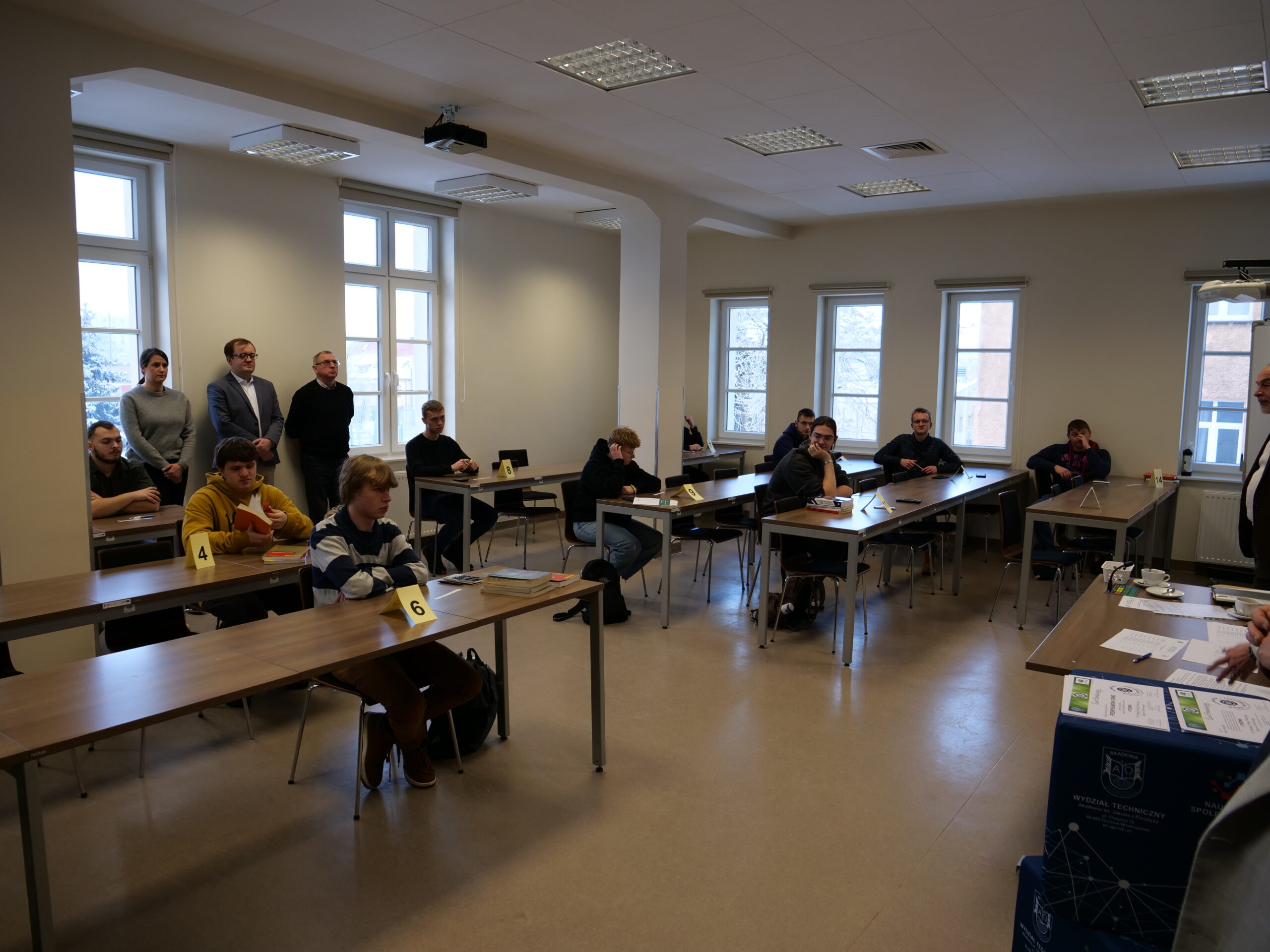 Uczestnicy olimpiady siedzą w ławkach. Trzy osoby stoją za nimi pod oknem.