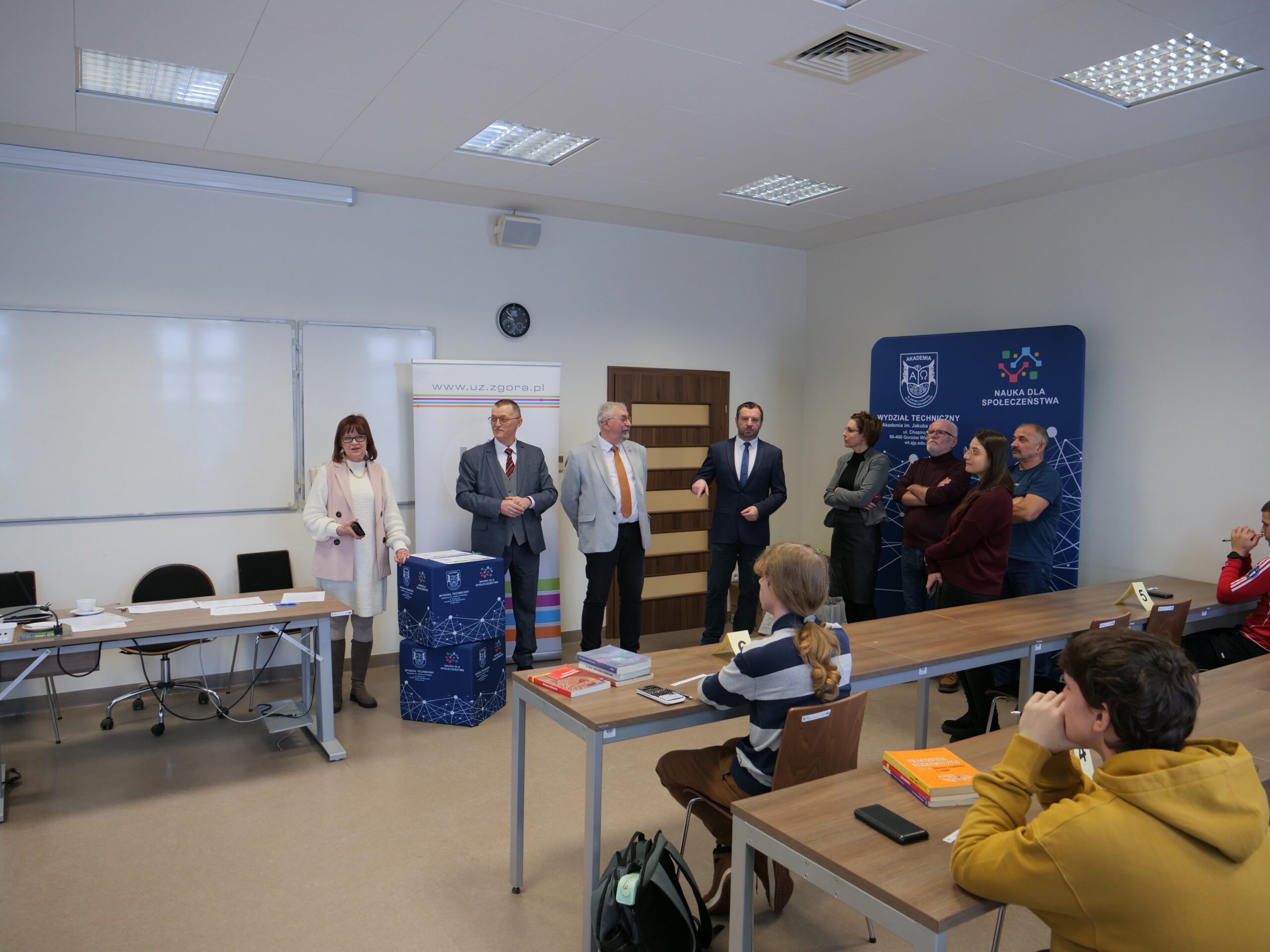 Uczestnicy olimpiady siedzą w ławkach, przed nimi stoją organizatorzy - osiem osób.