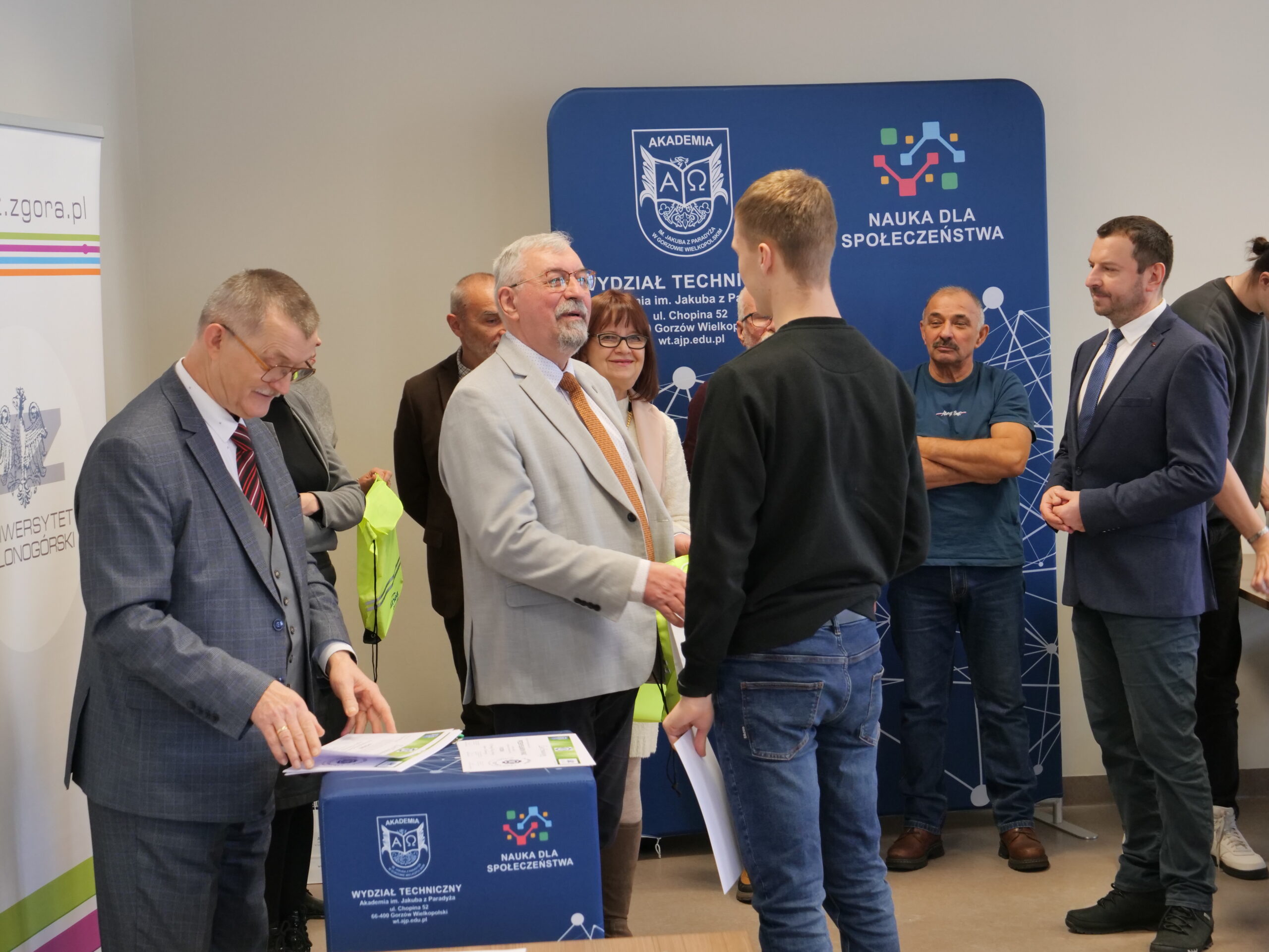 Uczestnik olimpiady odbiera od organizatora certyfikat, obok stoi grupa osób.