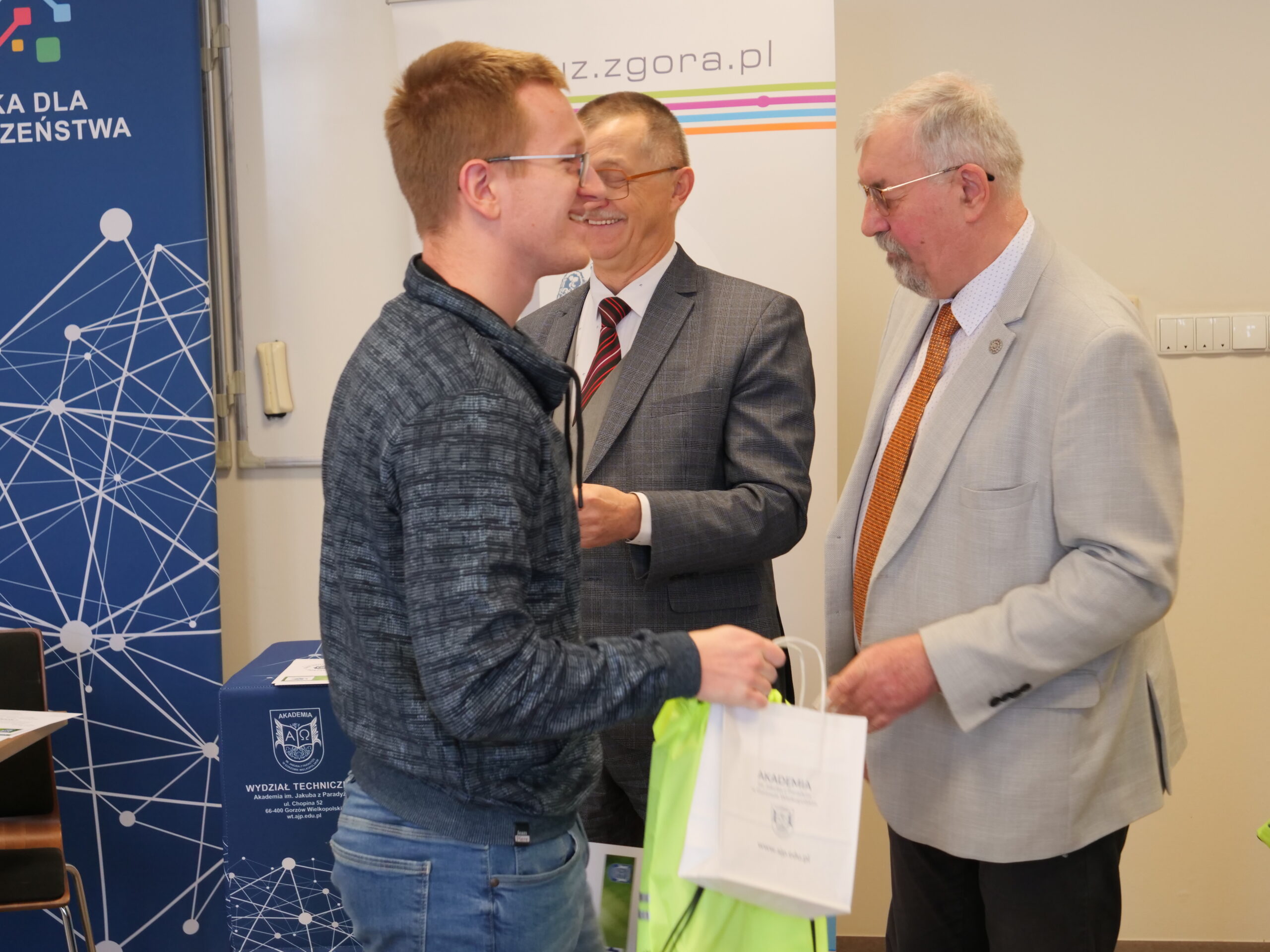 Uczestnik olimpiady odbiera od organizatora certyfikat, obok stoi trzeci mężczyzna z nagrodą w ręku.