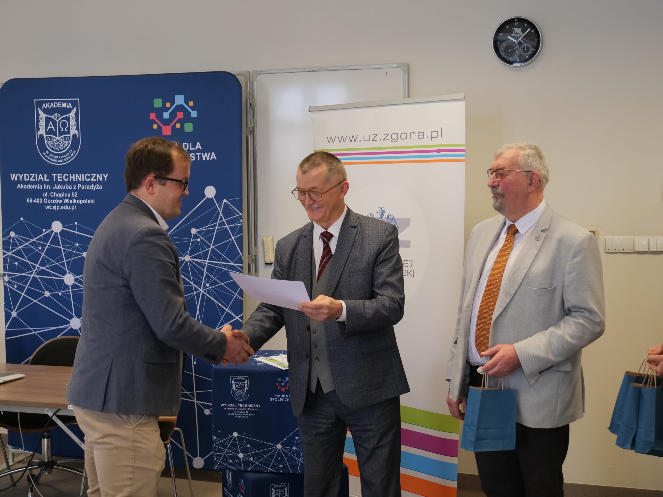 Uczestnik olimpiady odbiera od organizatora certyfikat, obok stoi trzeci mężczyzna z nagrodą w ręku.