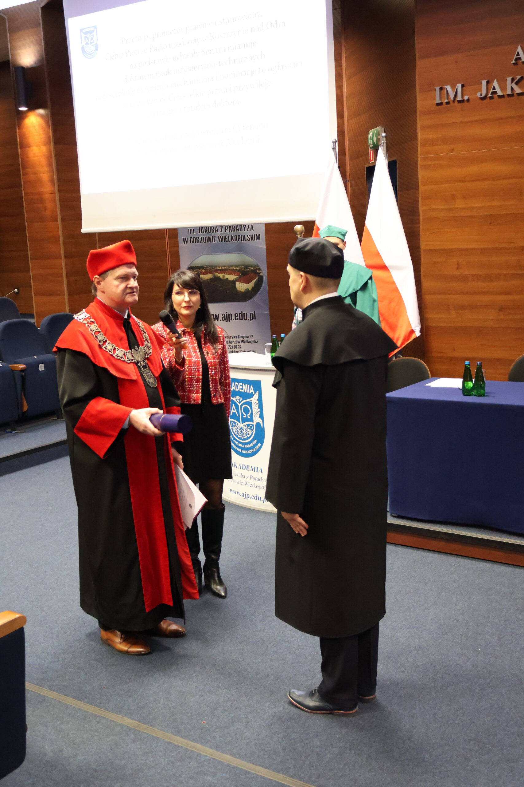 Prorektor wręcza Doktorowi dyplom, obok stoi kobieta i trzyma mikrofon