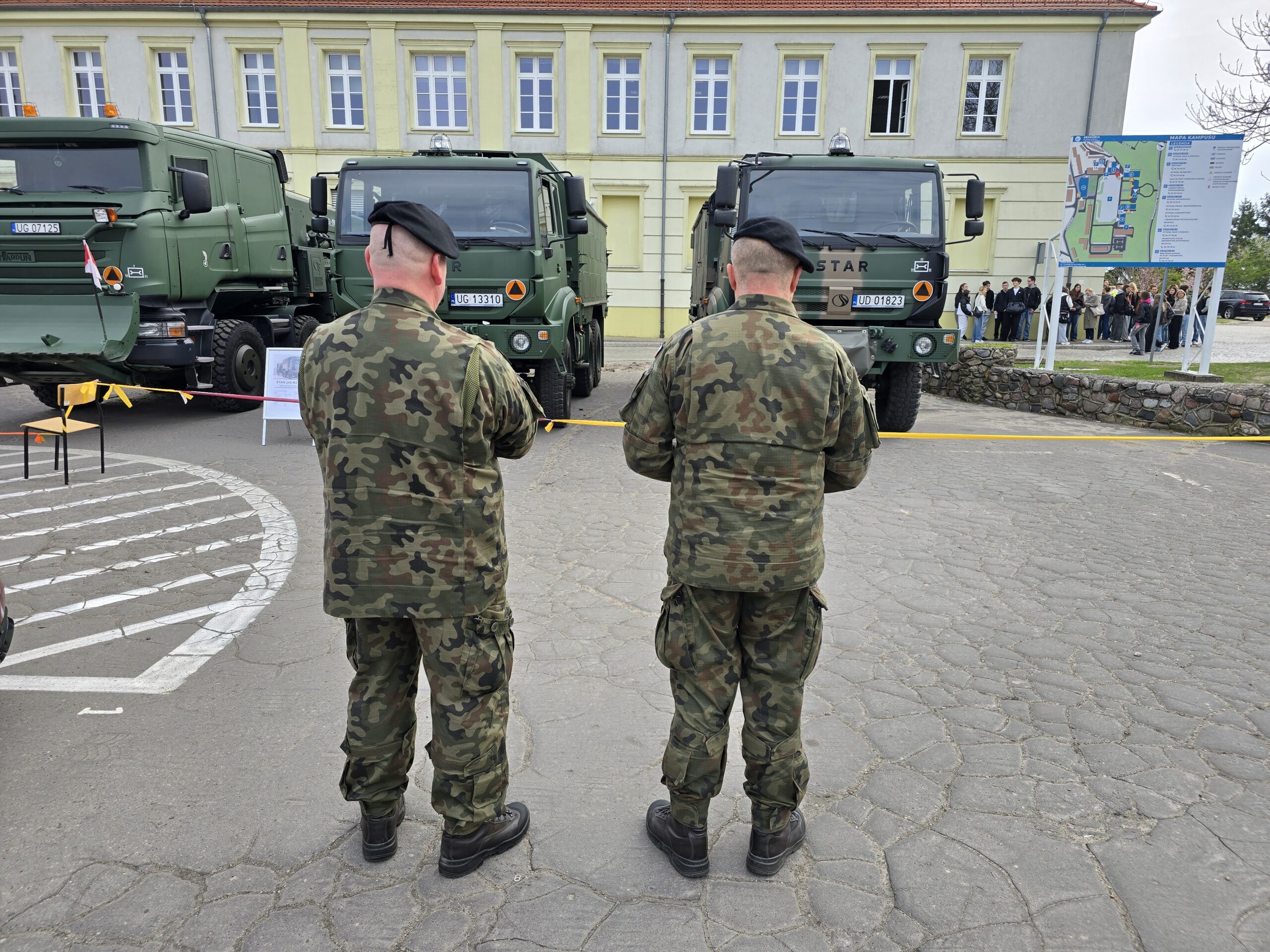 Dwóch żołnierzy stoi tyłem przy wojskowych samochodach.