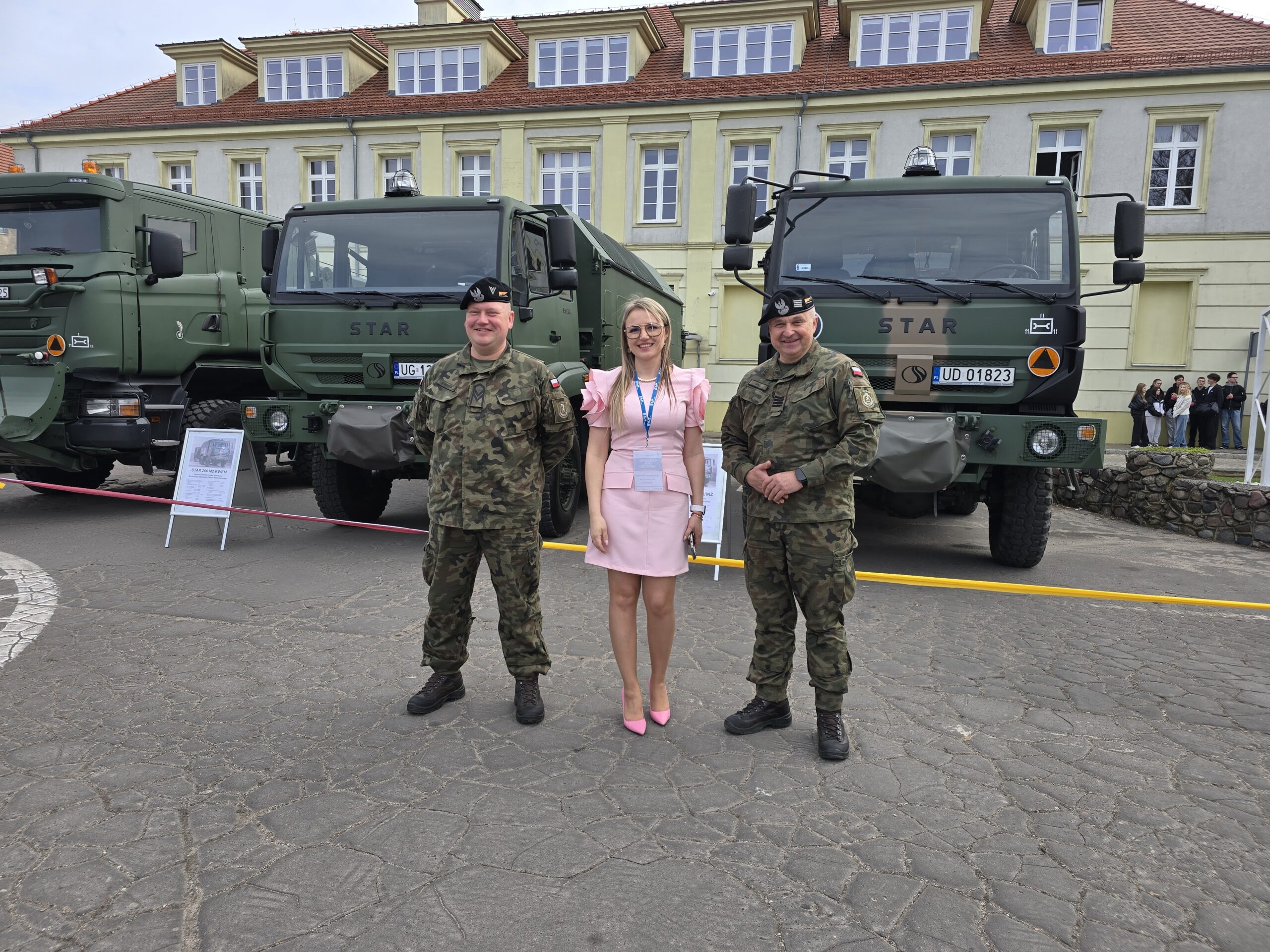 Pracownik AJP stoi z dwoma żołnierzami przy wojskowych wozach.