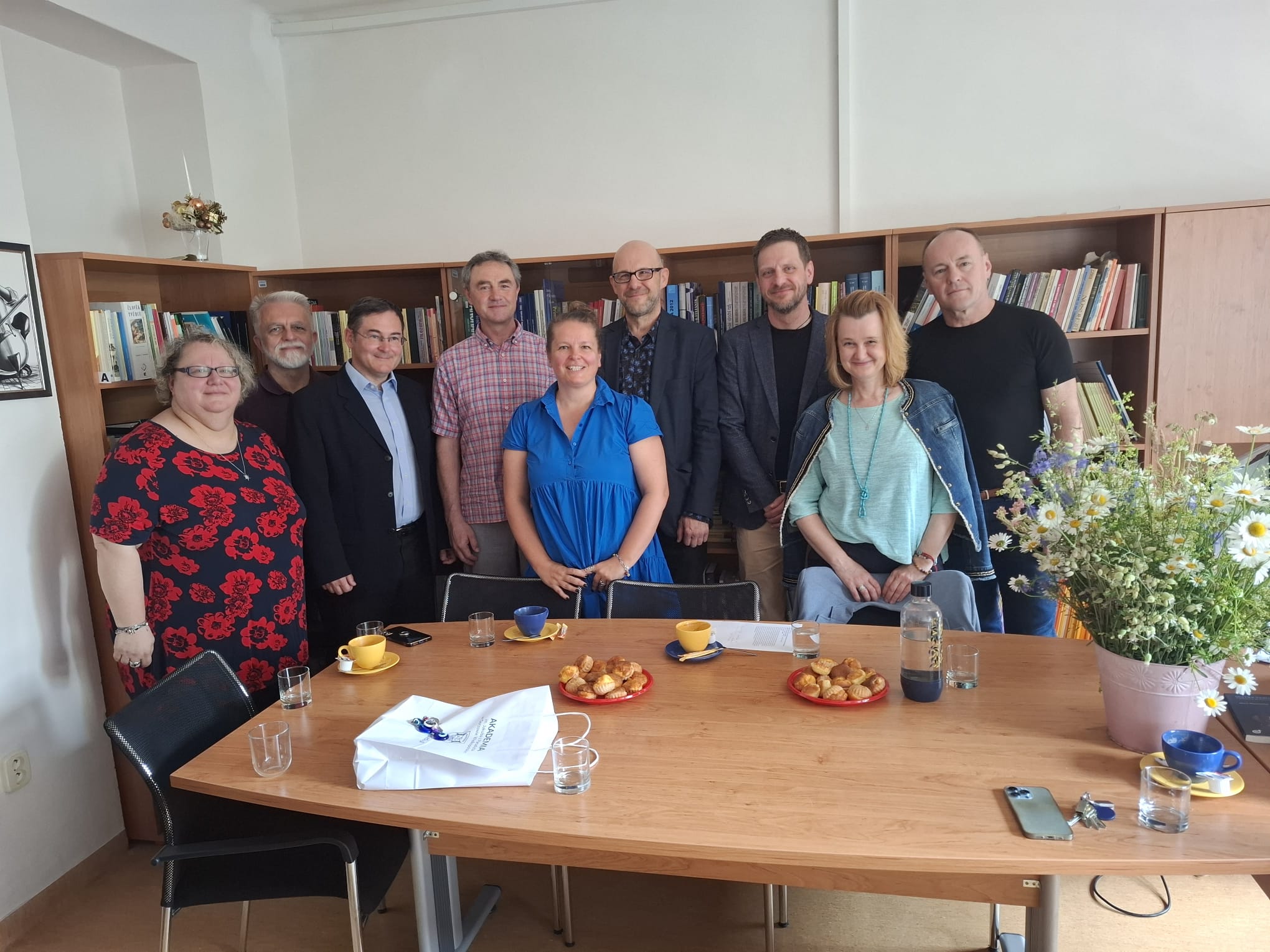 Pracownicy Katedry Filozofii i Nauk Politycznych oraz Wydziału Prawa i Bezpieczeństwa AJP są gromadzeni za stołem w gabinecie uniwersyteckim, za nimi znajdują się regały z książkami. Na stole stoją filiżanki, talerzyki z przekąskami, szklanki z wodą i bukiet polnych kwiatów. Od lewej: dr Radoslava Brhlíková w czarnej bluzce w czerwone kwiaty; prof. Ján Zozulak z siwą brodą; doc. Tomáš Pružinec w garniturze; doc. Radoslav Ivančík w koszuli w kratkę; dr Mária Kočnerová w niebieskiej sukience; dr Tomasz Marcinkowski w ciemnej marynarce; dr Juliusz Sikorski w granatowej marynarce; doc. Tatiana Tokolyová w jasnoniebieskim swetrze z kurtką; dr Andrzej Skwarski w czarnej koszulce. Wszyscy uśmiechają się przyjaźnie.