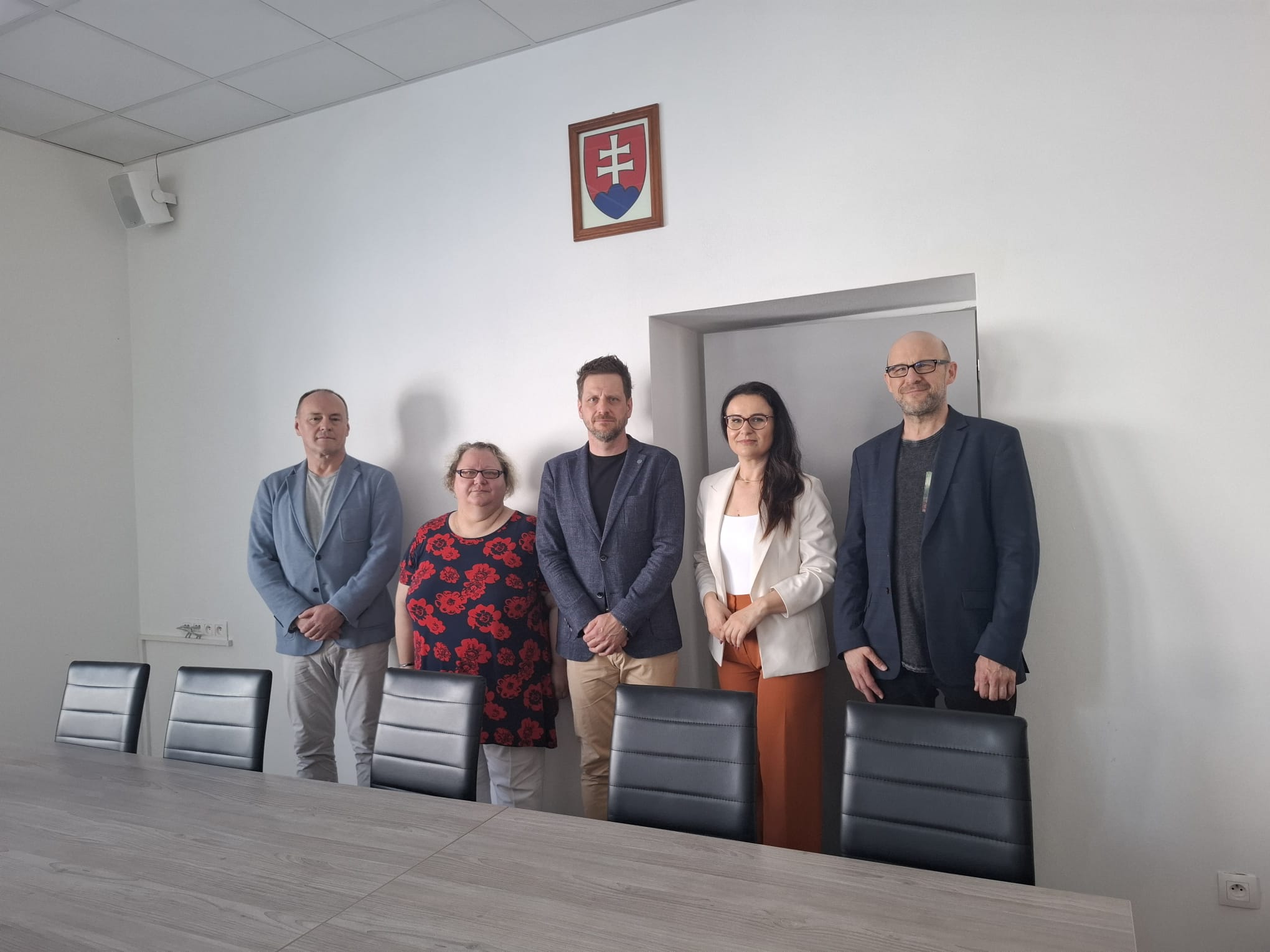 Dwie kobiety i trzech mężczyzn stoi obok pustego stołu konferencyjnego w jasnej sali, nad nimi wisi godło Słowacji. Od lewej: dr Andrzej Skwarski w jasnoniebieskiej marynarce; dr Radoslava Brhlíková w czarnej bluzce w duże czerwone kwiaty; dr Juliusz Sikorski w ciemnej marynarce; dr Daša Nováčiková w białej marynarce i pomarańczowych spodniach; dr Tomasz Marcinkowski w ciemnej marynarce i czarnej koszulce. Wszyscy patrzą w obiektyw i subtelnie się uśmiechają.