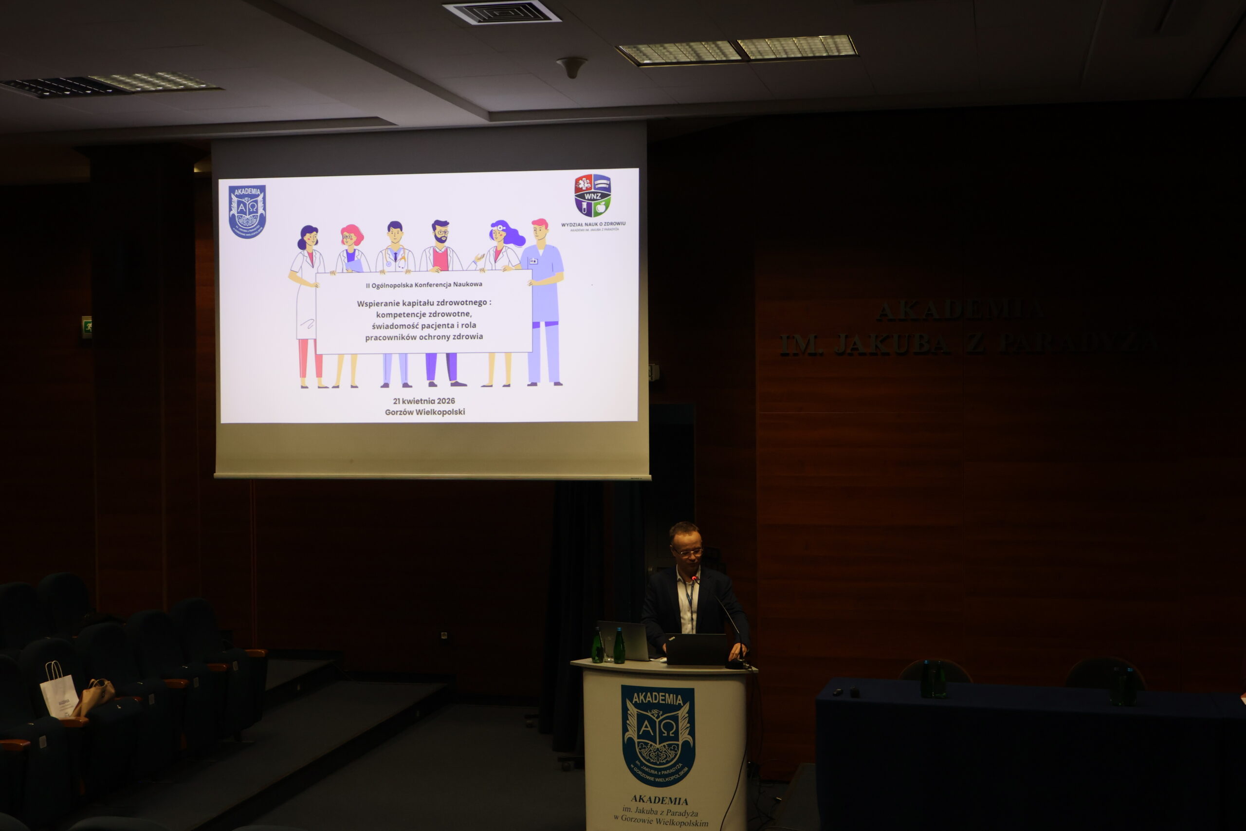 Prelegent stoi przy mównicy podczas II Ogólnopolskiej Konferencji Naukowej "Wspieranie kapitału zdrowotnego, kompetencje zdrowotne, świadomość pacjenta i rola pracowników ochrony zdrowia".