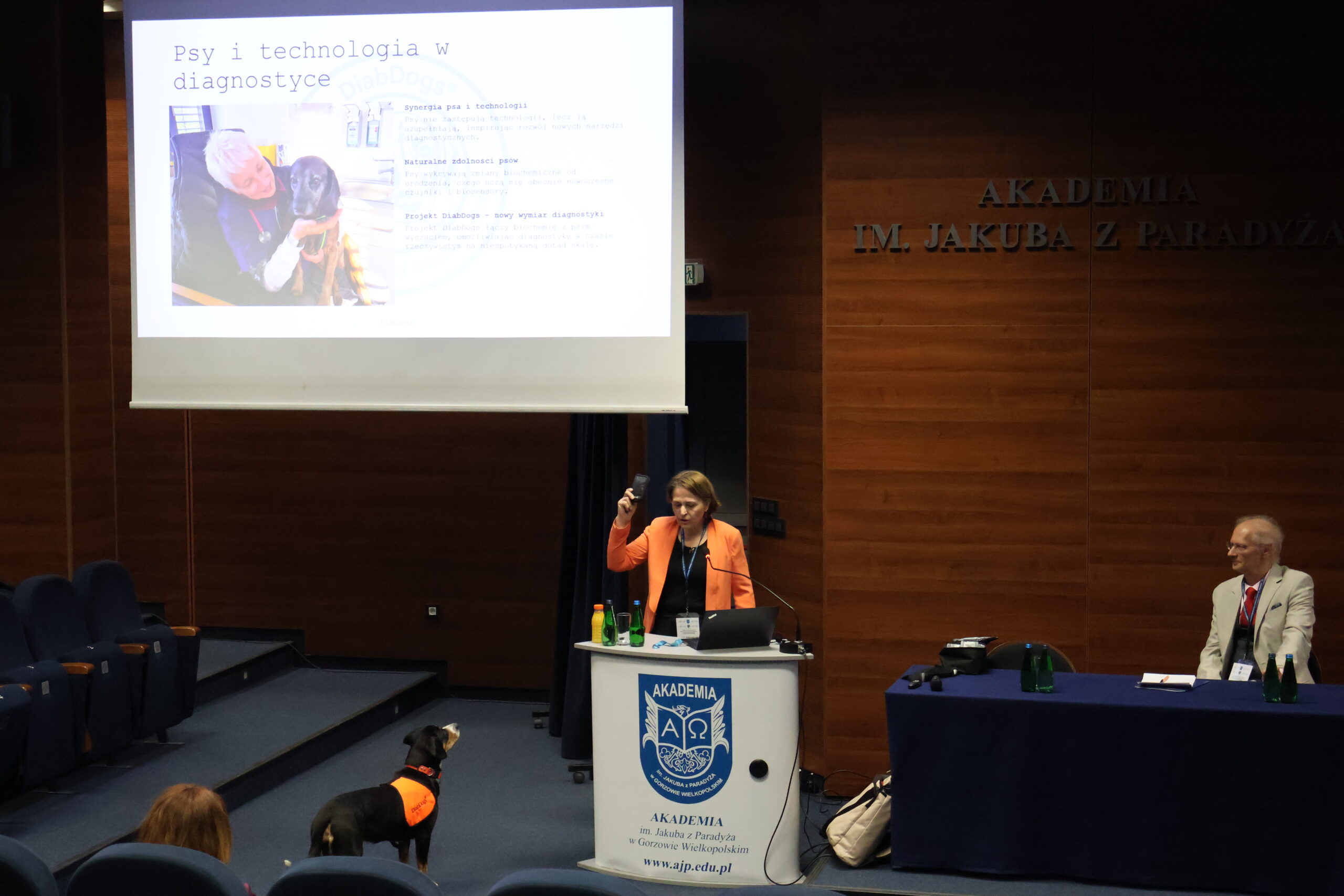 Prelegentka prezentująca temat Pies w diagnostyce cukrzycowej. Na slajdzie: Psy i technologia w diagnostyce. Obok mównicy znajduje się pies w ubraniu ratowniczym,