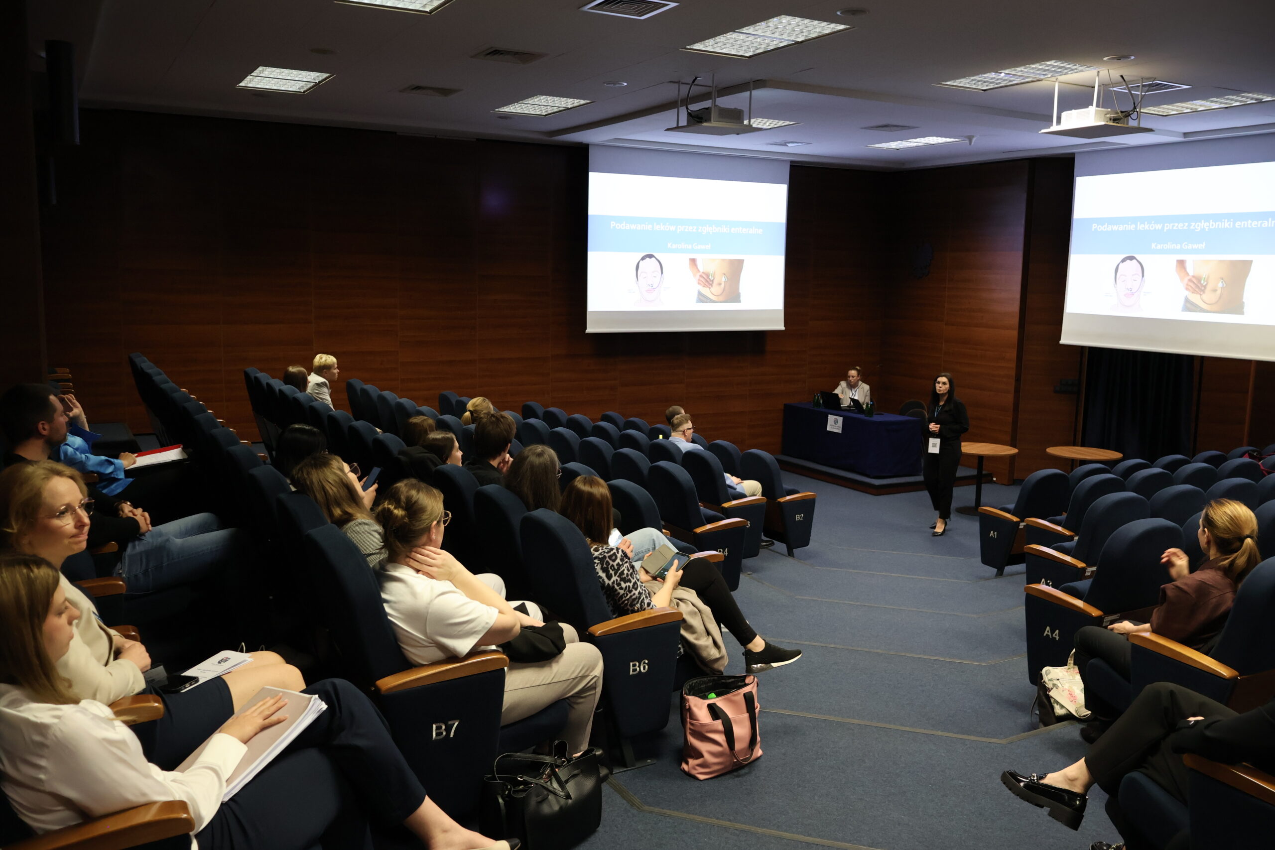 Kobieta znajduje się na środku auli prezentująca dla przybyłych temat Podawanie leków przez zgłębniki enteralne.