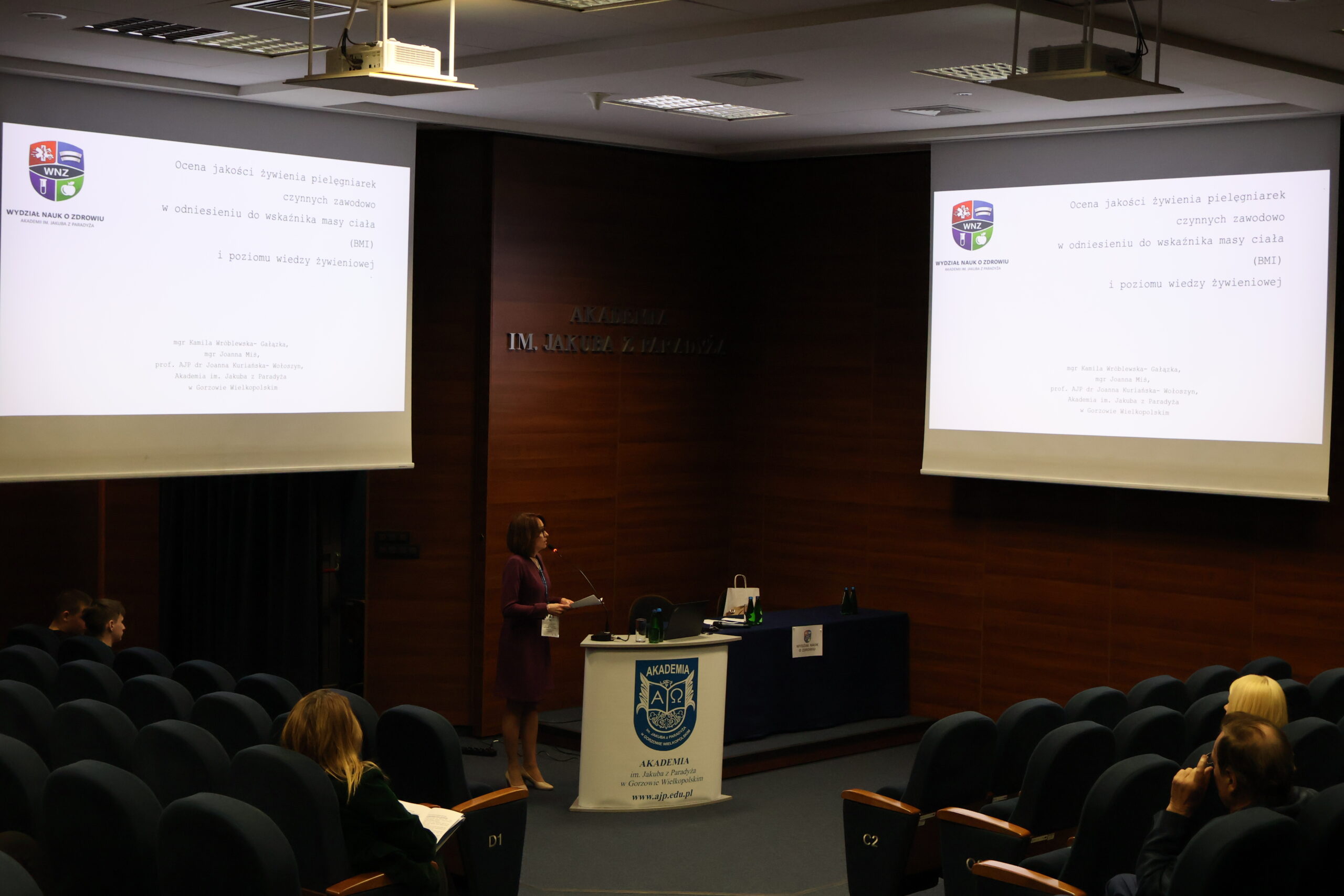 Na zdjęciu kobieta przemawiająca na konferencji patrząca na slajd i prezentująca swój temat pod tytułem "Ocena jakości żywienia pielęgniarek czynnych zawodowo w odniesieniu do wskaźnika masy ciała (BMI) i poziom wiedzy żywieniowej".